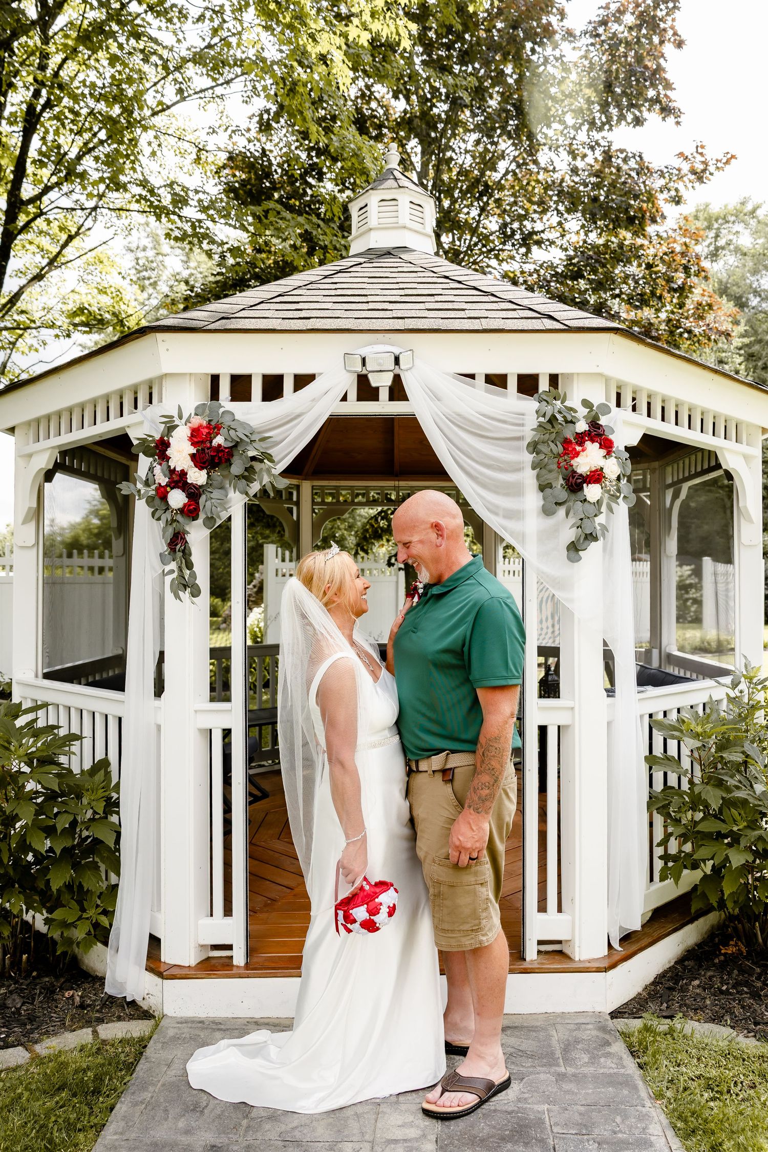 Bride and groom stand together in front of a white decorated gazebo with red and white floral arrangements.