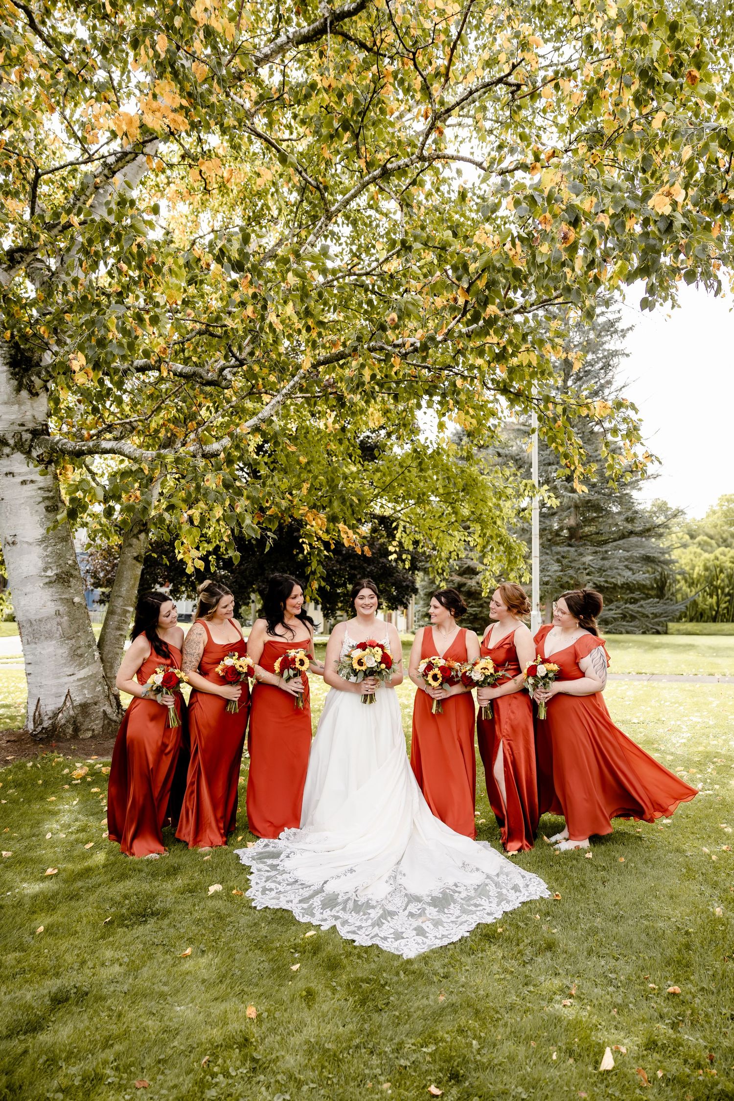 Bride in white gown with bridesmaids in red dresses holding sunflower bouquets under autumn tree on green lawn.