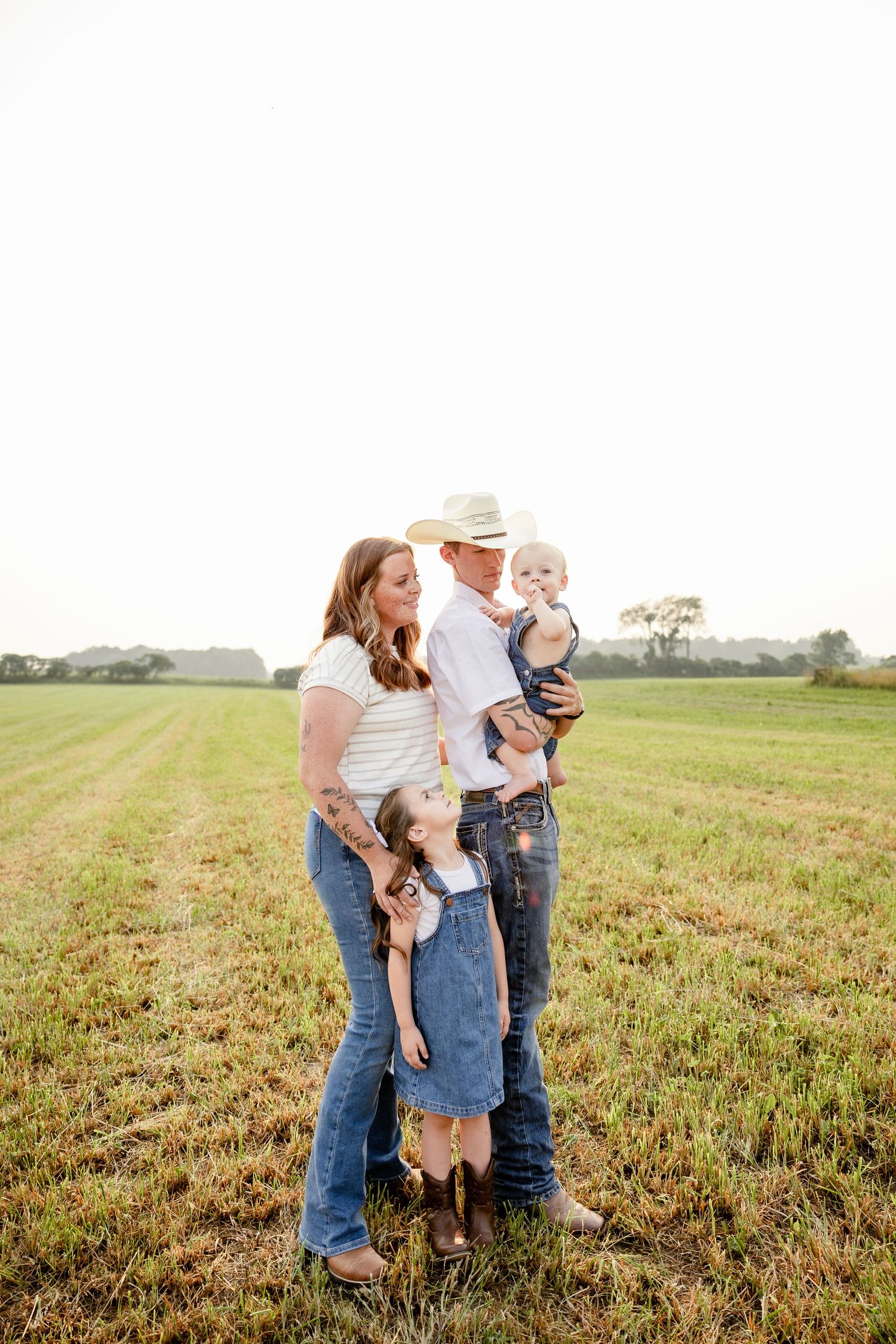 Family of four posing in a green field, parents in jeans and cowboy hats holding children on a sunny day.