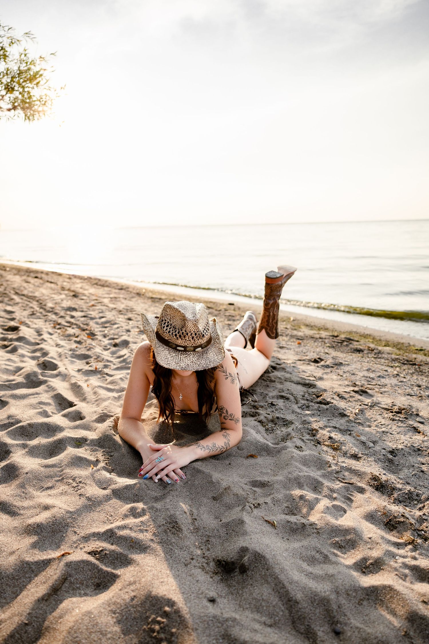 Woman in straw cowboy hat and boots lying on sandy beach near calm ocean at sunset.
