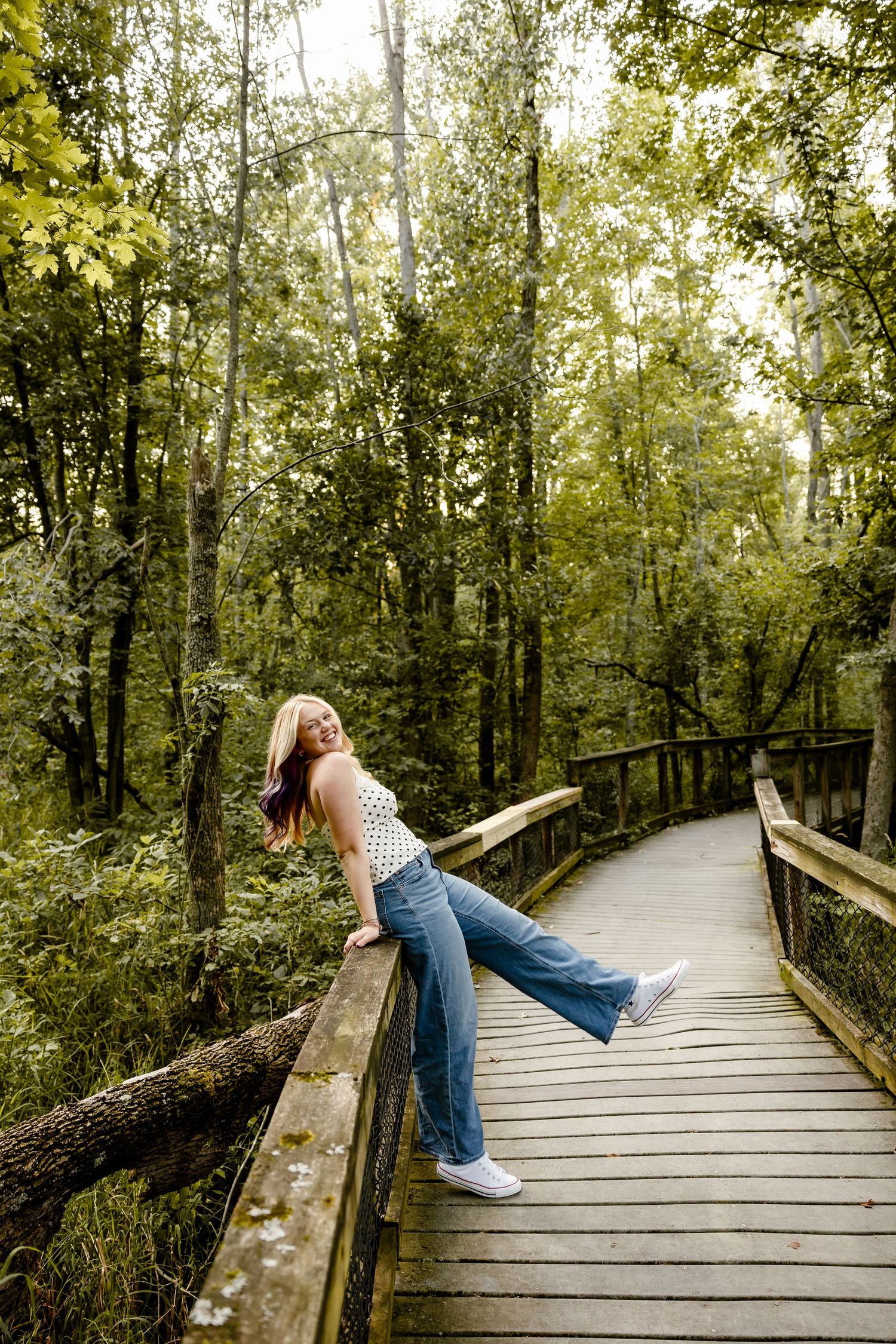 Young woman in flare jeans poses on wooden boardwalk bridge surrounded by lush green forest trees.
