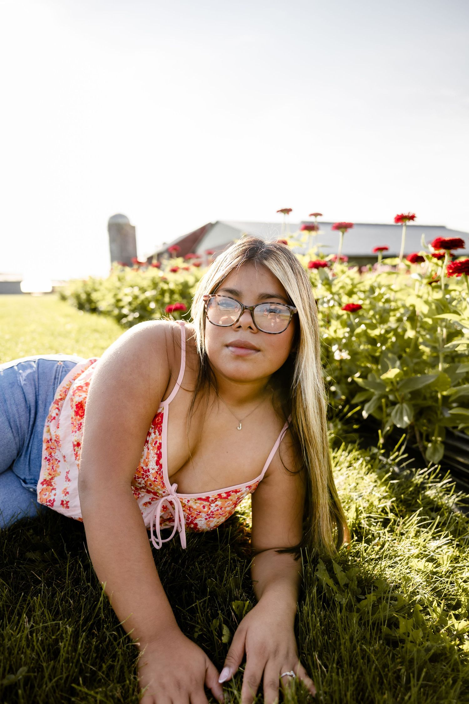 Young woman with glasses lying in green grass at a flower farm with red blooms and barn in background.