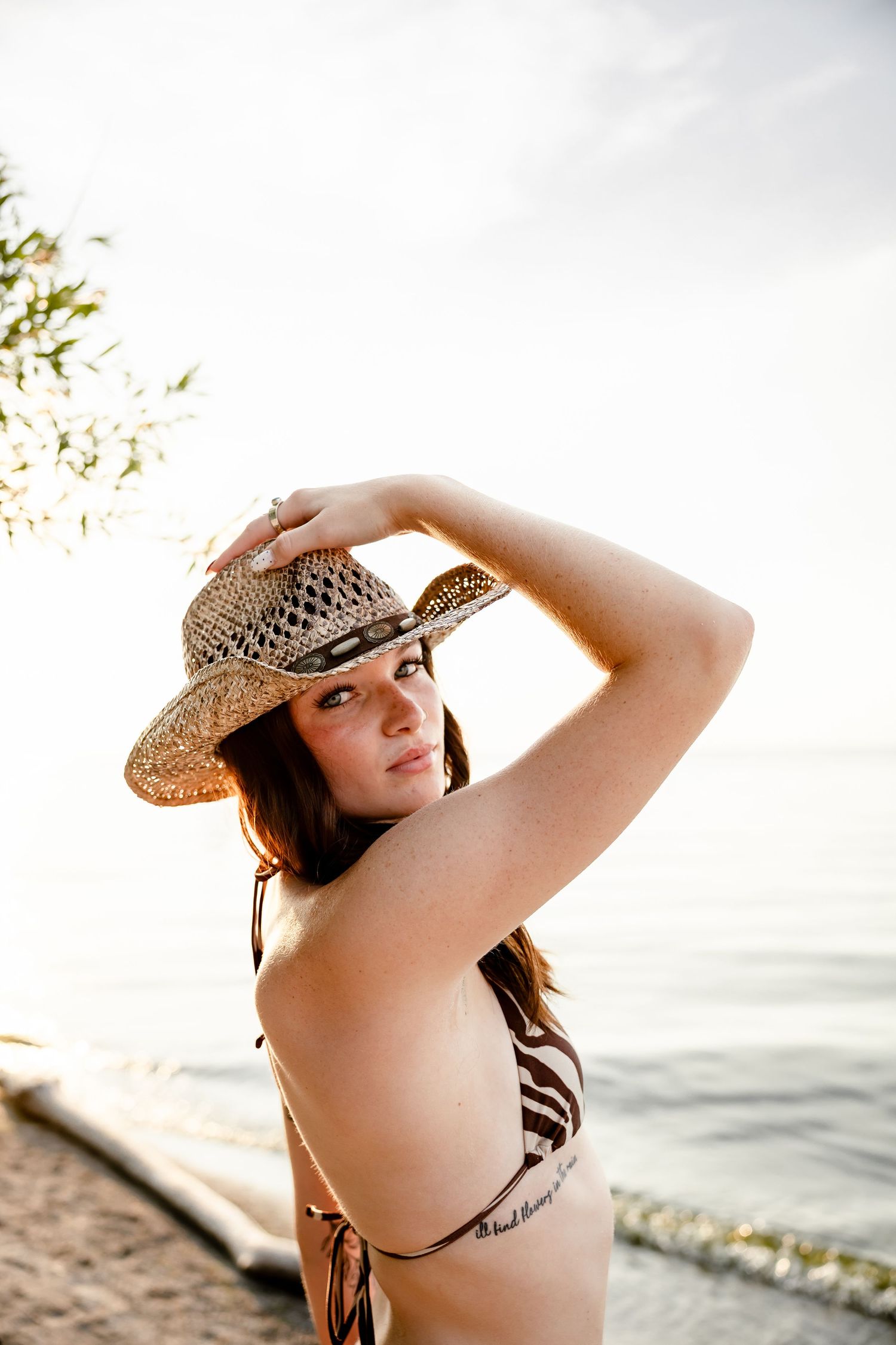 Young woman in striped bikini and straw hat posing at beach during golden sunset hour.