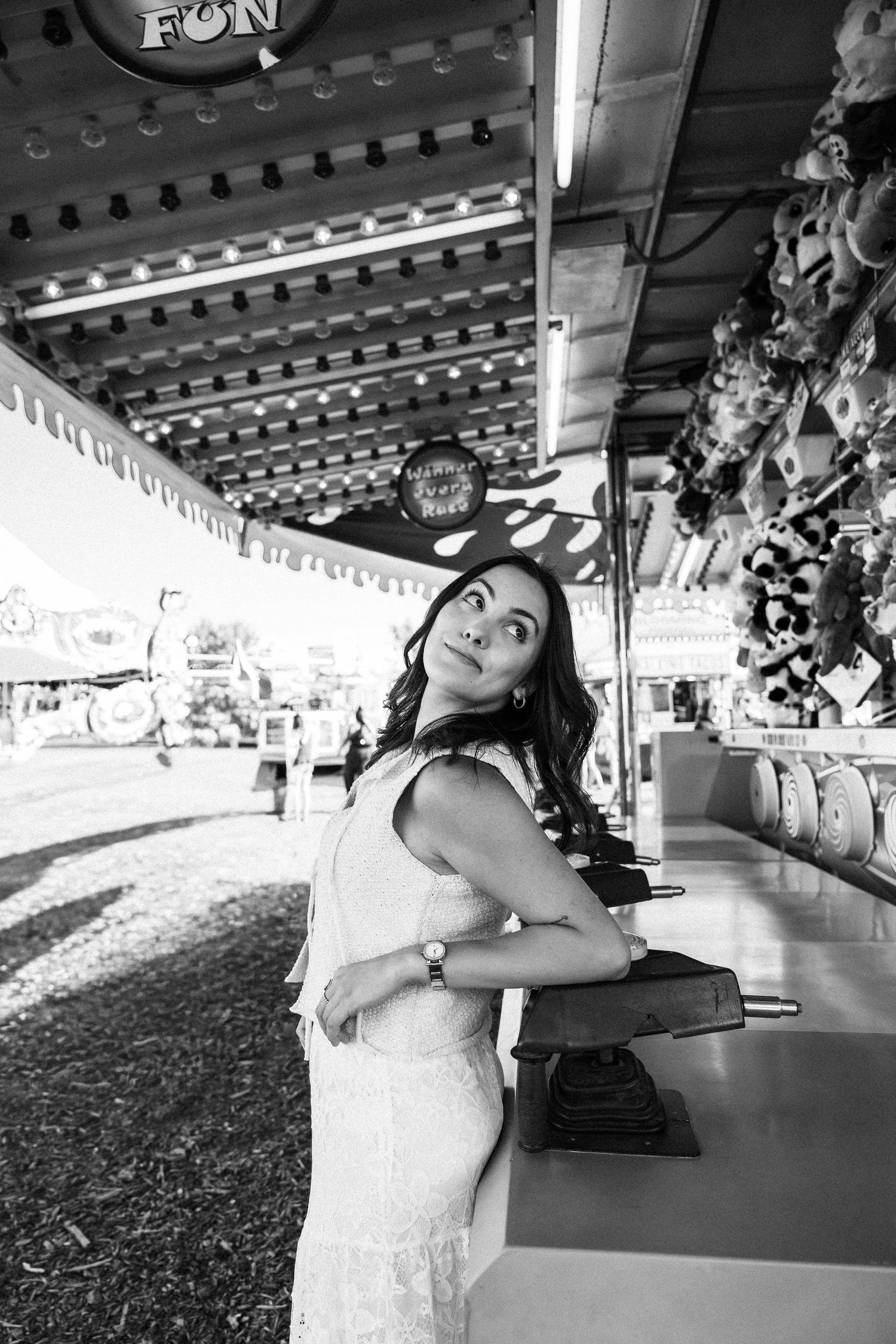 Black and white photo of a young woman in a white dress posing at a carnival game booth.
