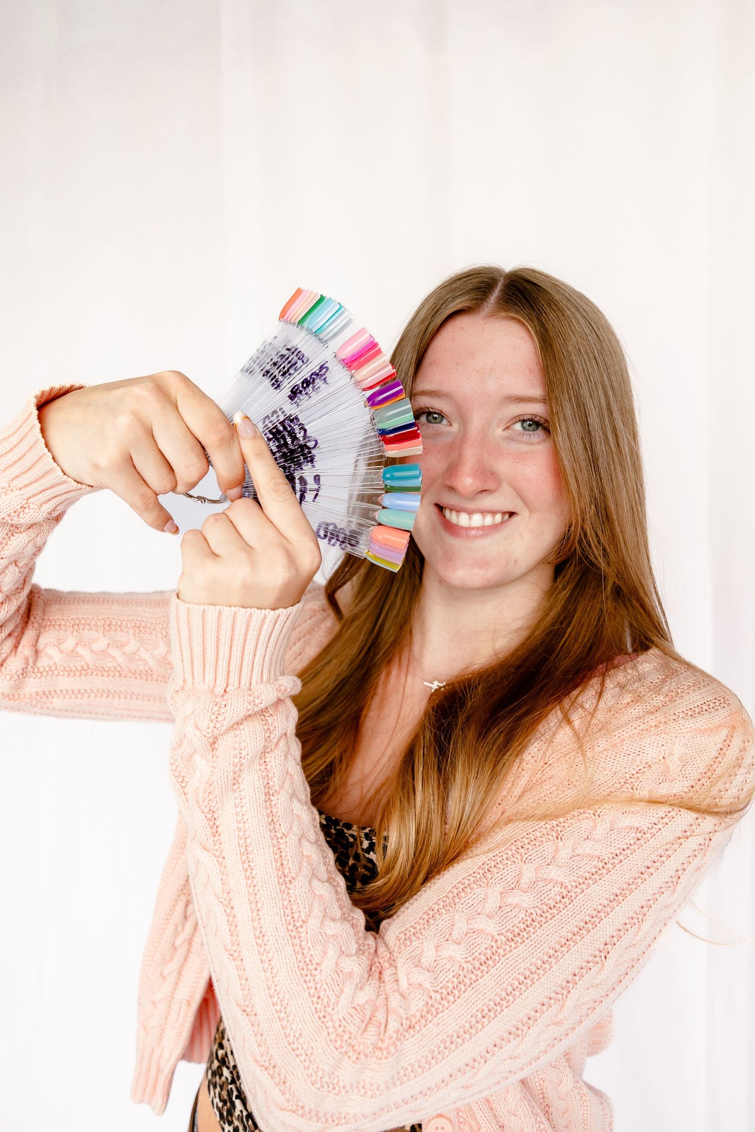 Smiling young woman in pink sweater holding colorful nail polish swatch fan in front of her face.