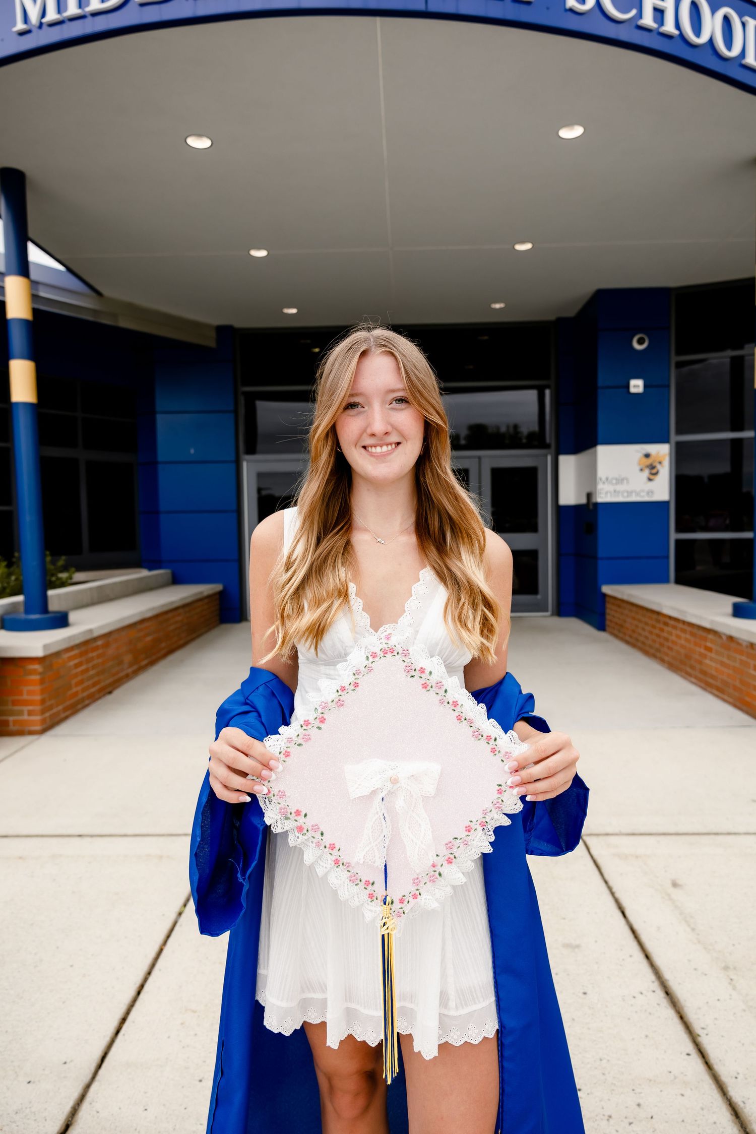 Smiling graduate in blue gown holds decorated cap outside school entrance.