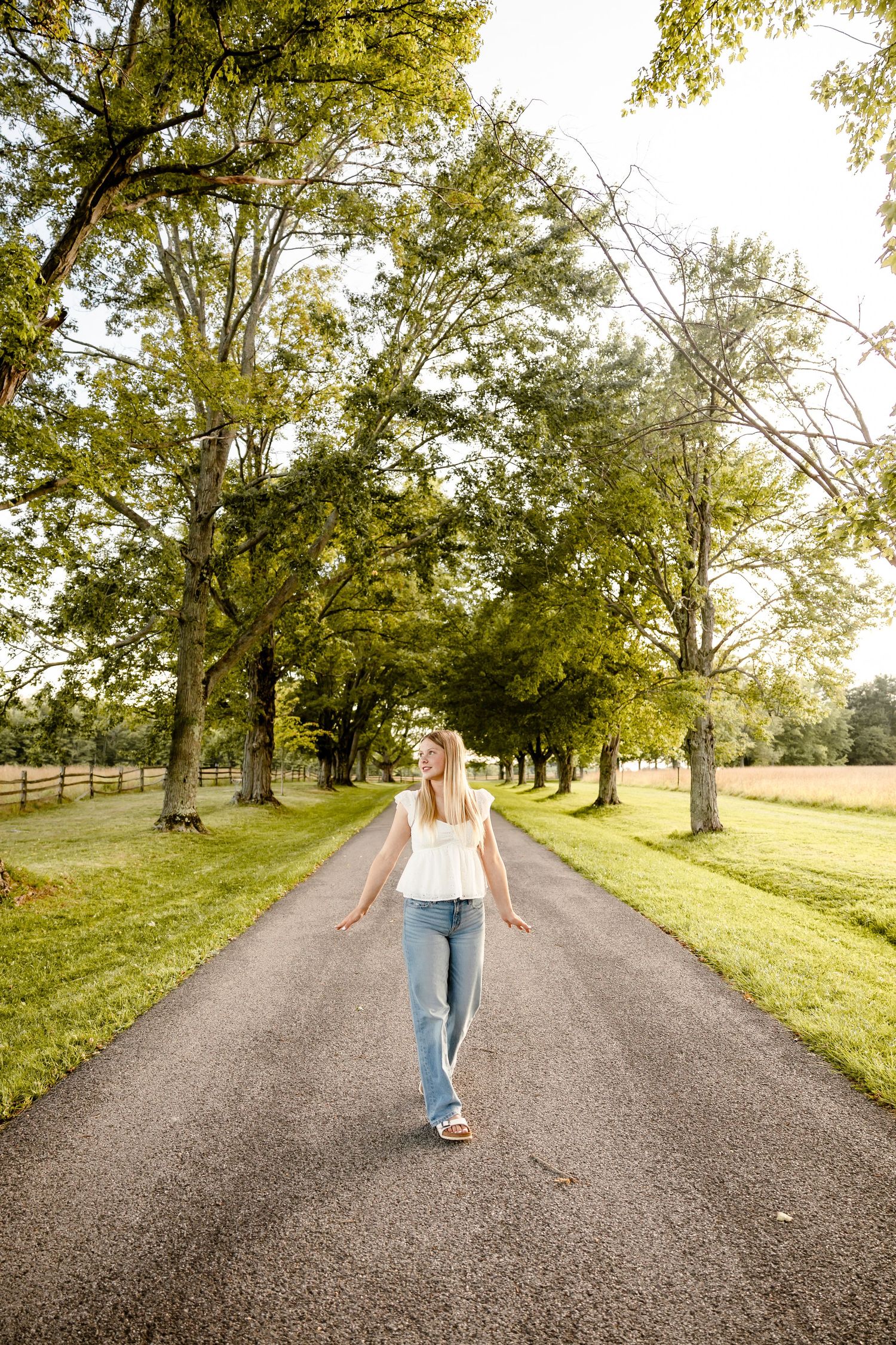 A young woman in white top and jeans walks down a tree-lined country road on a sunny day.