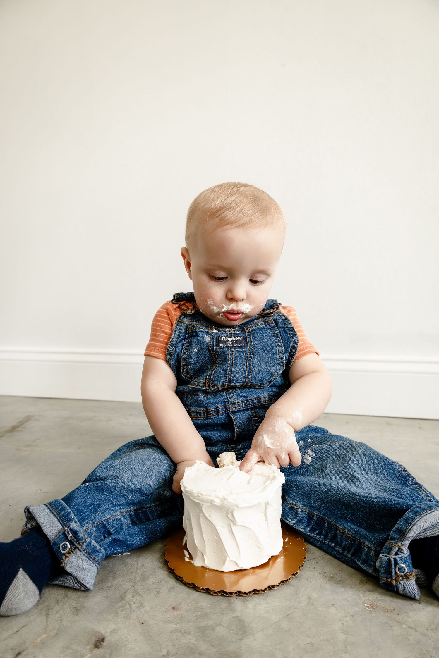 Toddler in denim overalls smashing a white frosted cake on floor during first birthday cake smash photo session.