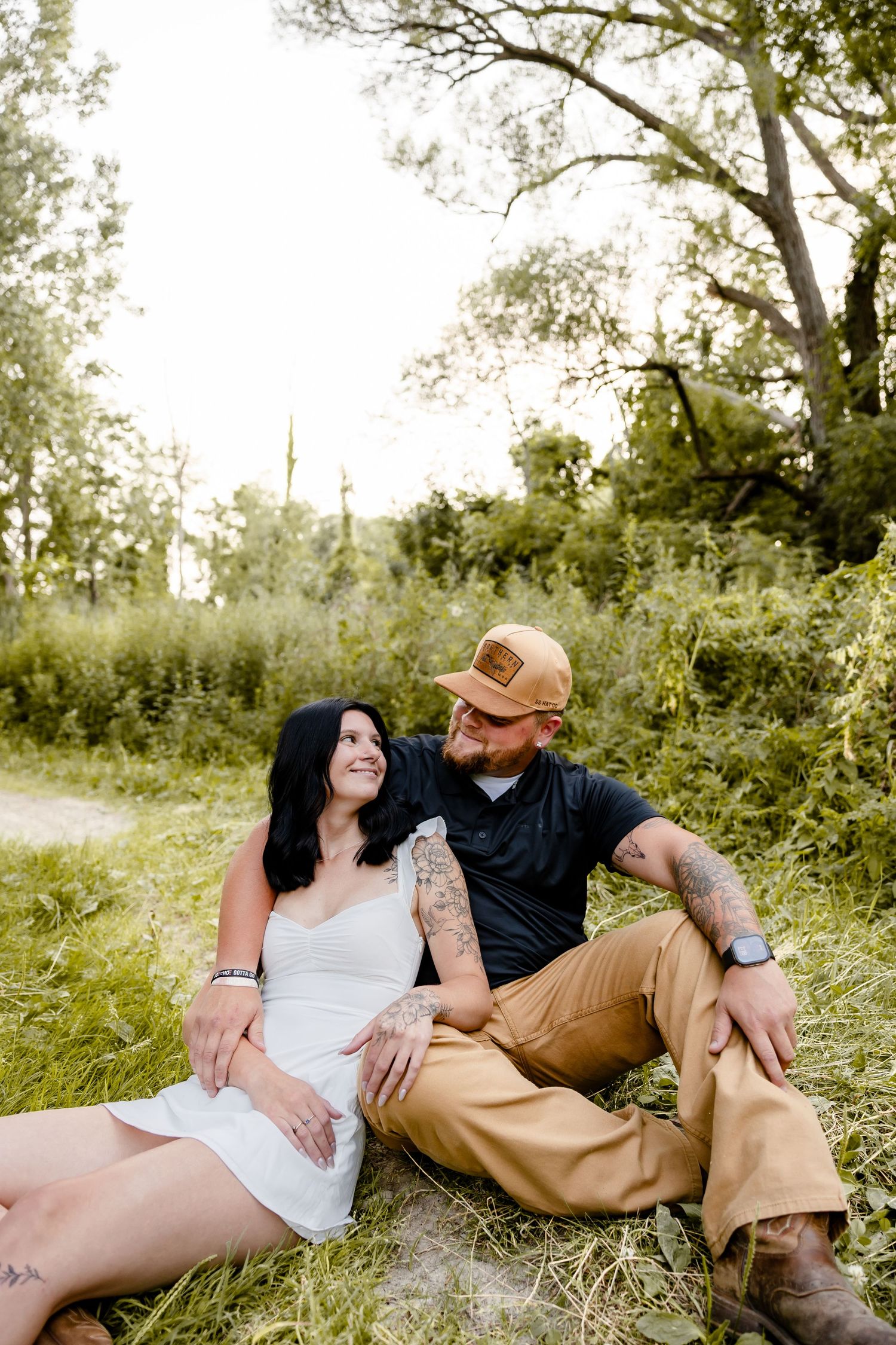 Couple with tattoos sitting together in a lush green field, smiling at each other, woman in white dress, man in cap.