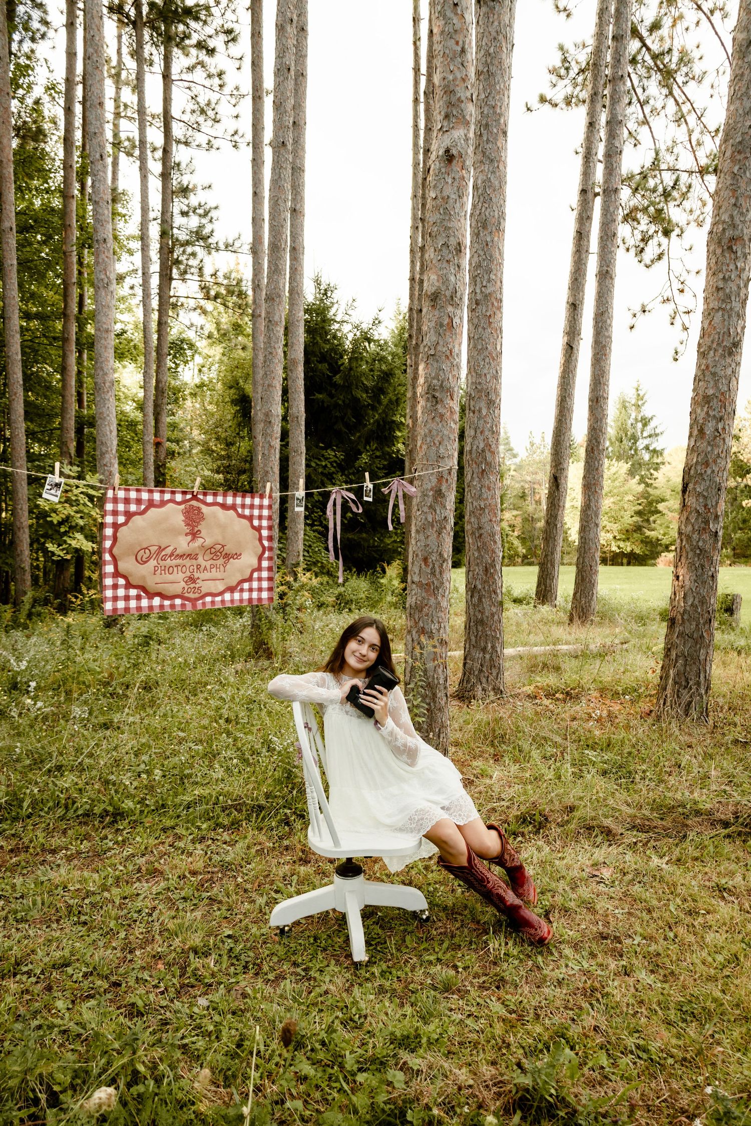 Young woman in white dress and cowboy boots relaxing in white chair among tall pine trees in a rustic outdoor setting.