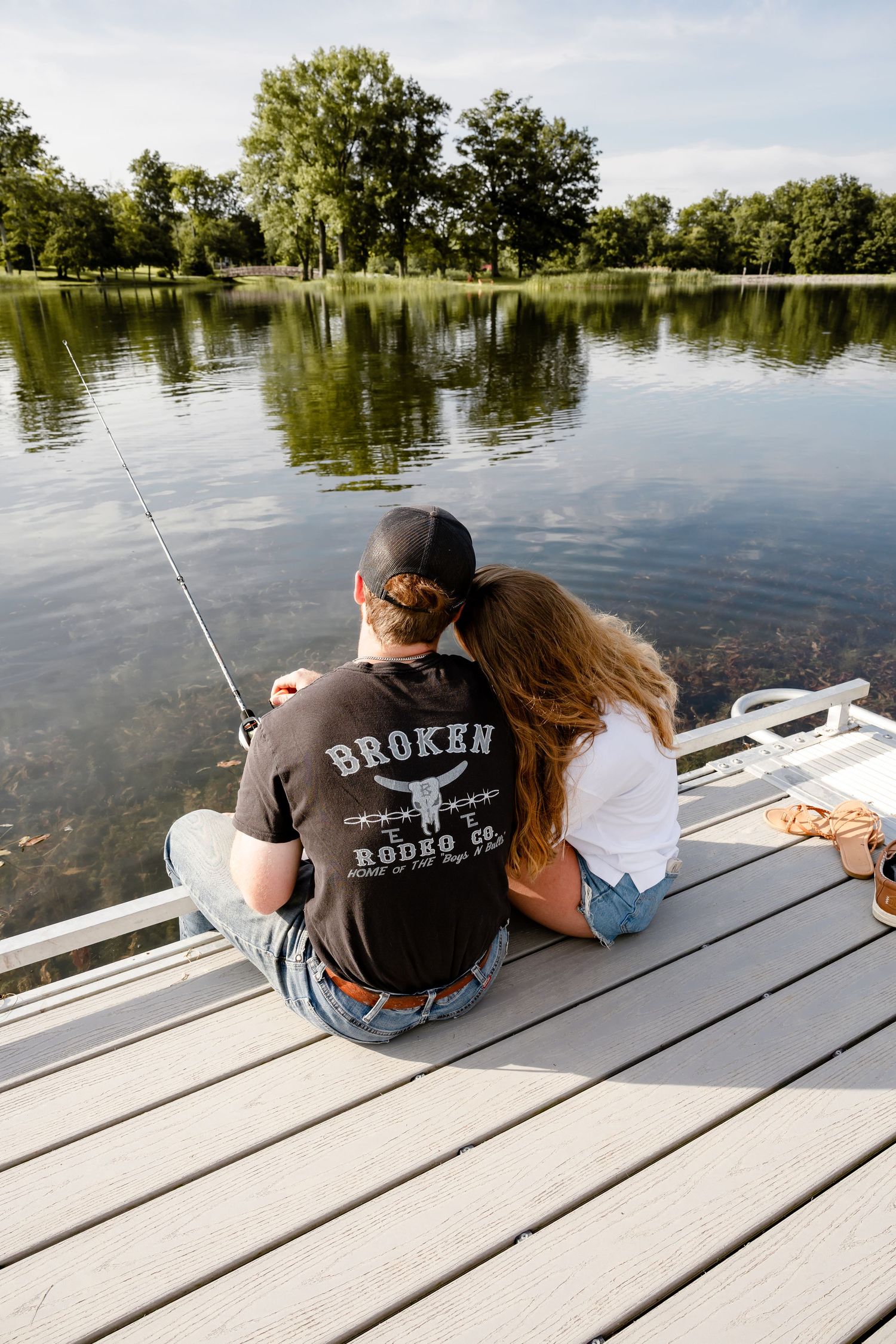 Couple sitting together on a dock fishing by a calm reflective lake surrounded by lush green trees.