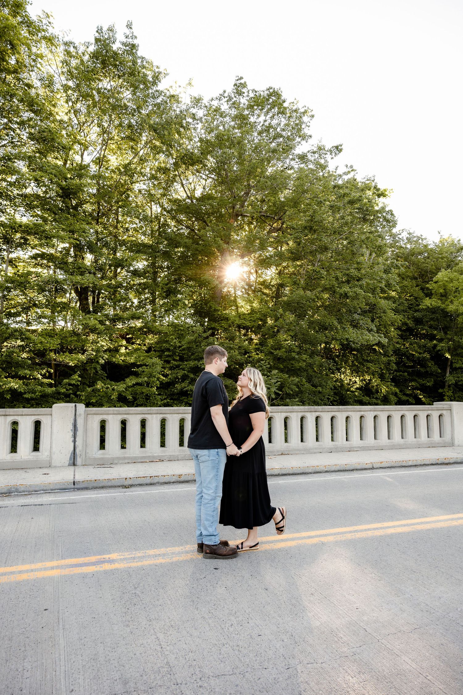 Couple holding hands on a bridge at golden hour, surrounded by lush green trees with sunlight streaming through.