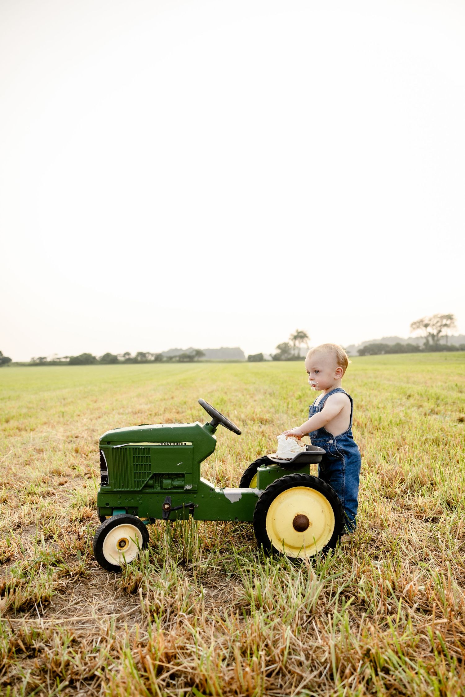 Toddler in denim overalls riding a green John Deere toy tractor in an open grassy field.