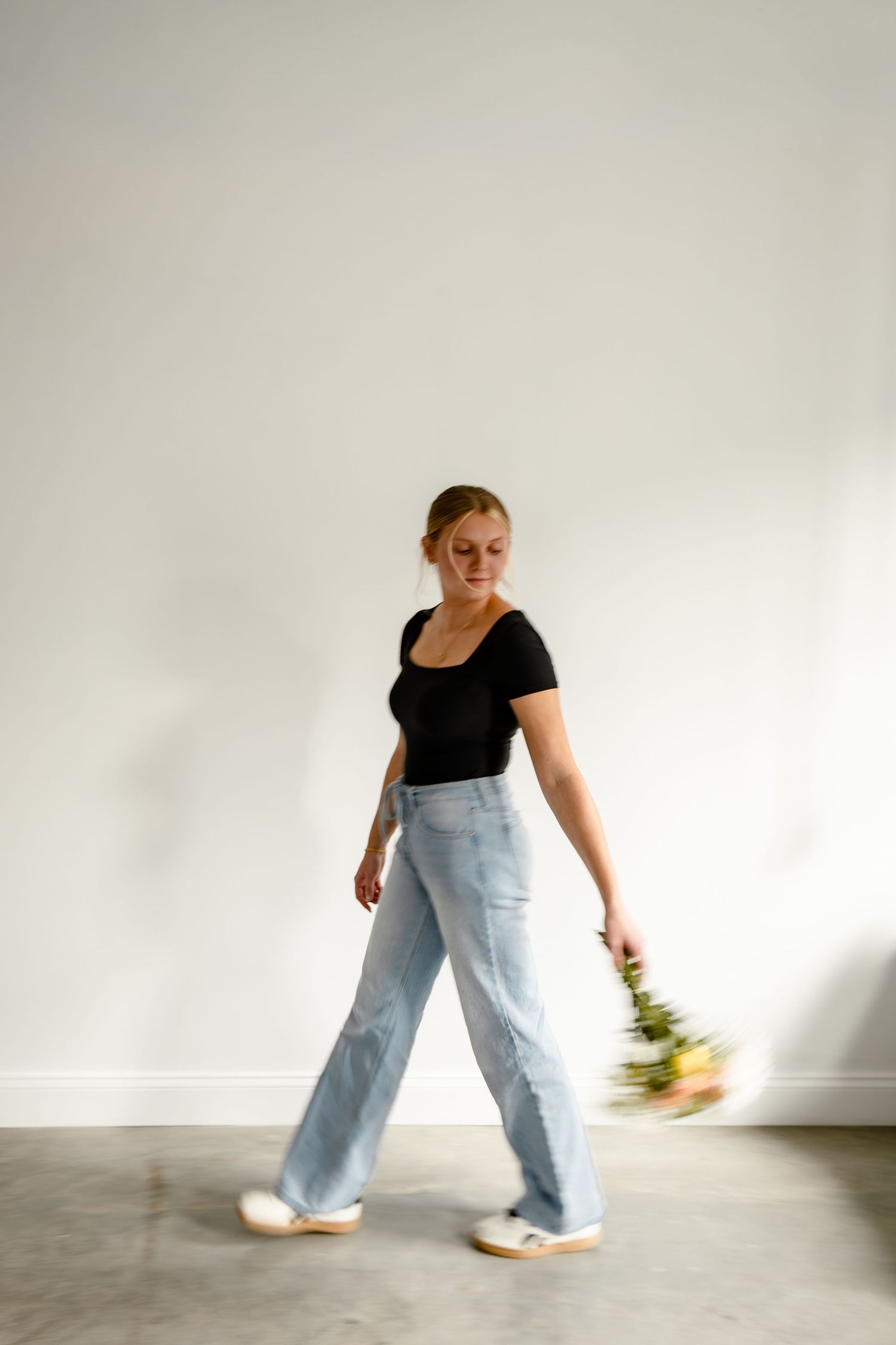 Woman in black top and light blue wide-leg jeans walking with flowers against a white wall.
