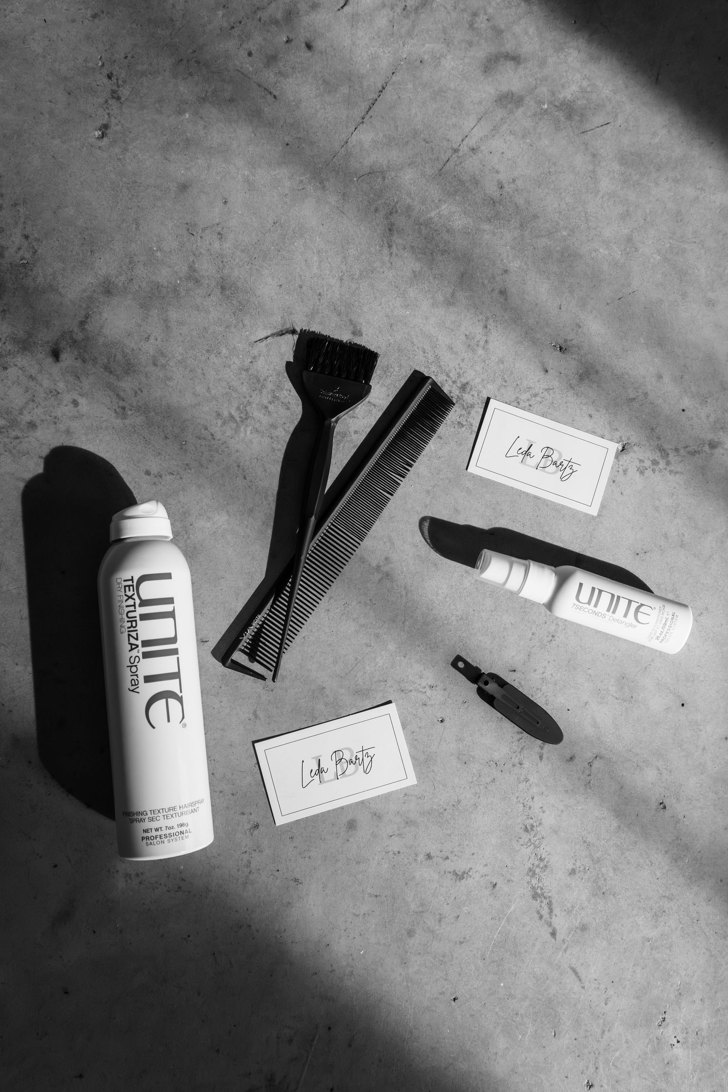 Black and white flatlay of Unite hair products, a comb, dye brush, clips, and Lob Boss business cards on concrete.