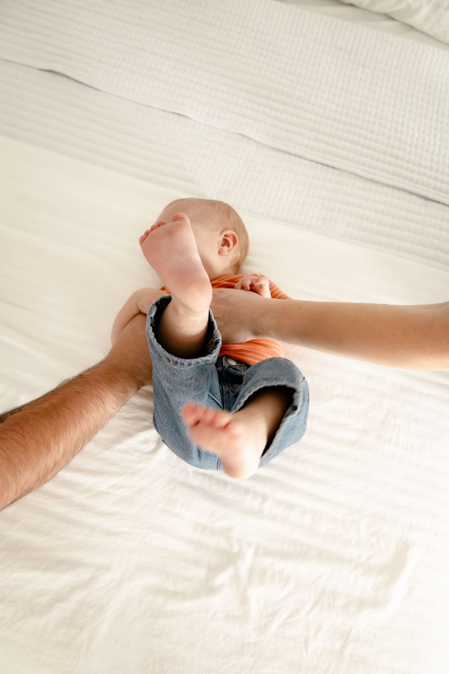 Baby in denim overalls lying on white bed with parent's hand nearby, barefoot infant relaxing on soft bedding.