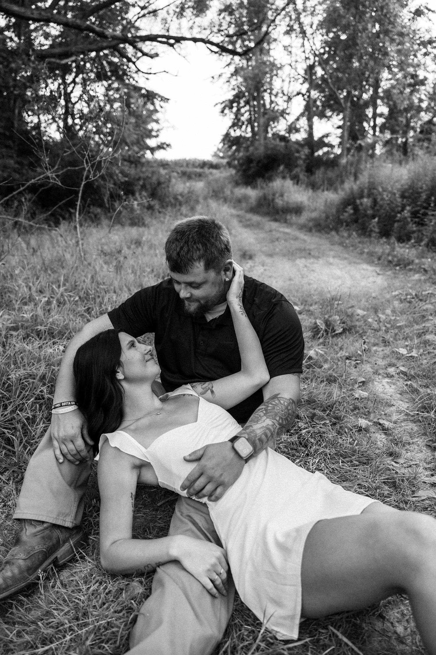 Black and white photo of a romantic couple gazing at each other, woman lying in grass, man leaning over her outdoors.