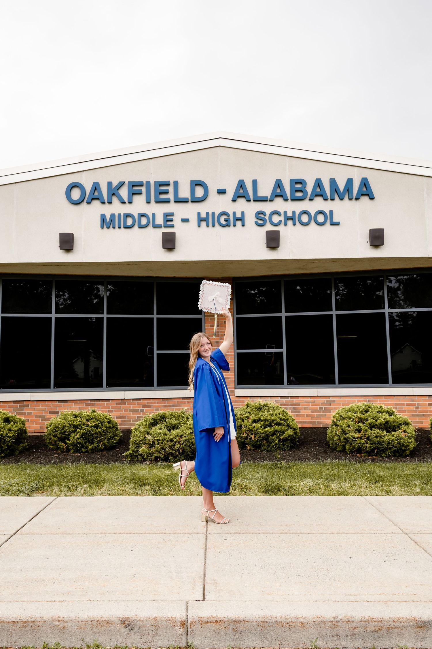 Graduate in blue cap and gown celebrates in front of Oakfield-Alabama Middle-High School building.