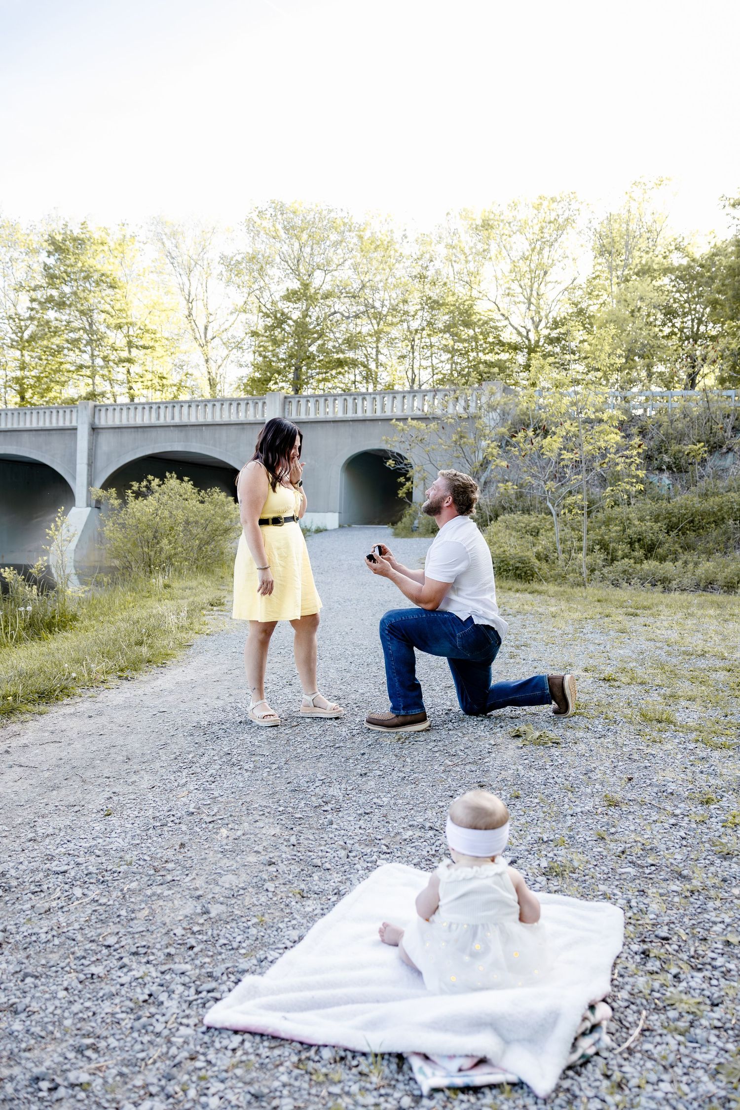 Man proposing on one knee to woman in yellow dress outdoors, baby on blanket nearby, bridge and trees in background.