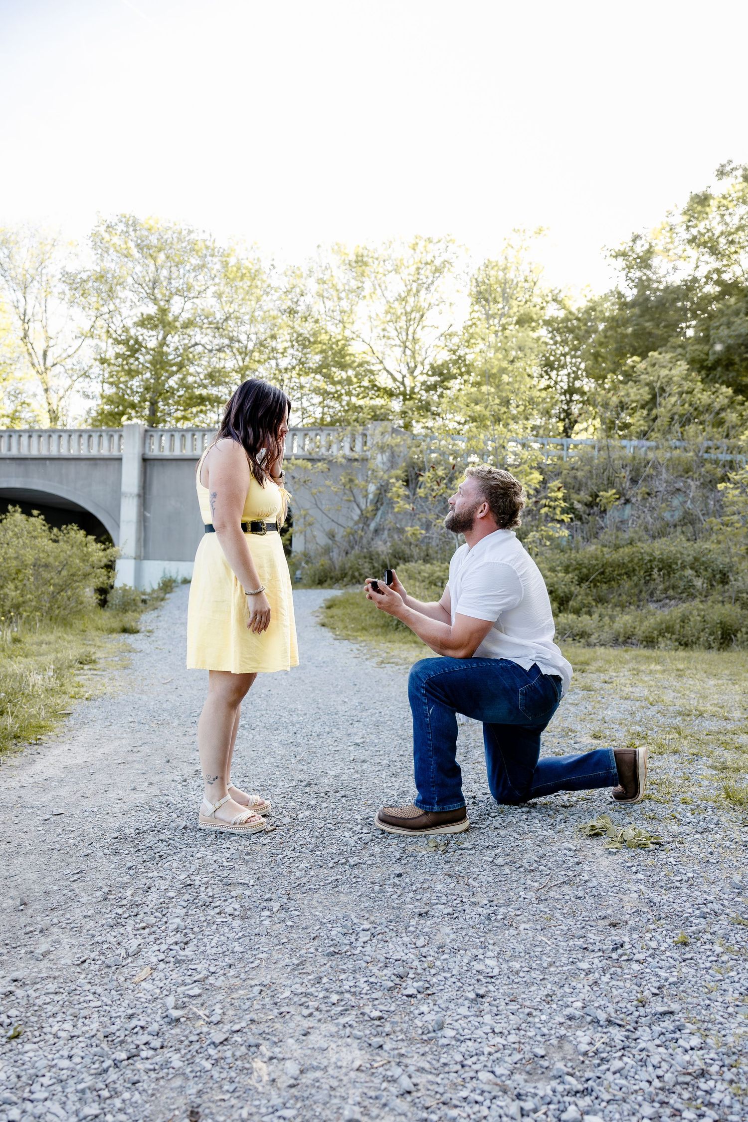 Man proposing on one knee to woman in yellow dress on gravel path with bridge and trees in background.