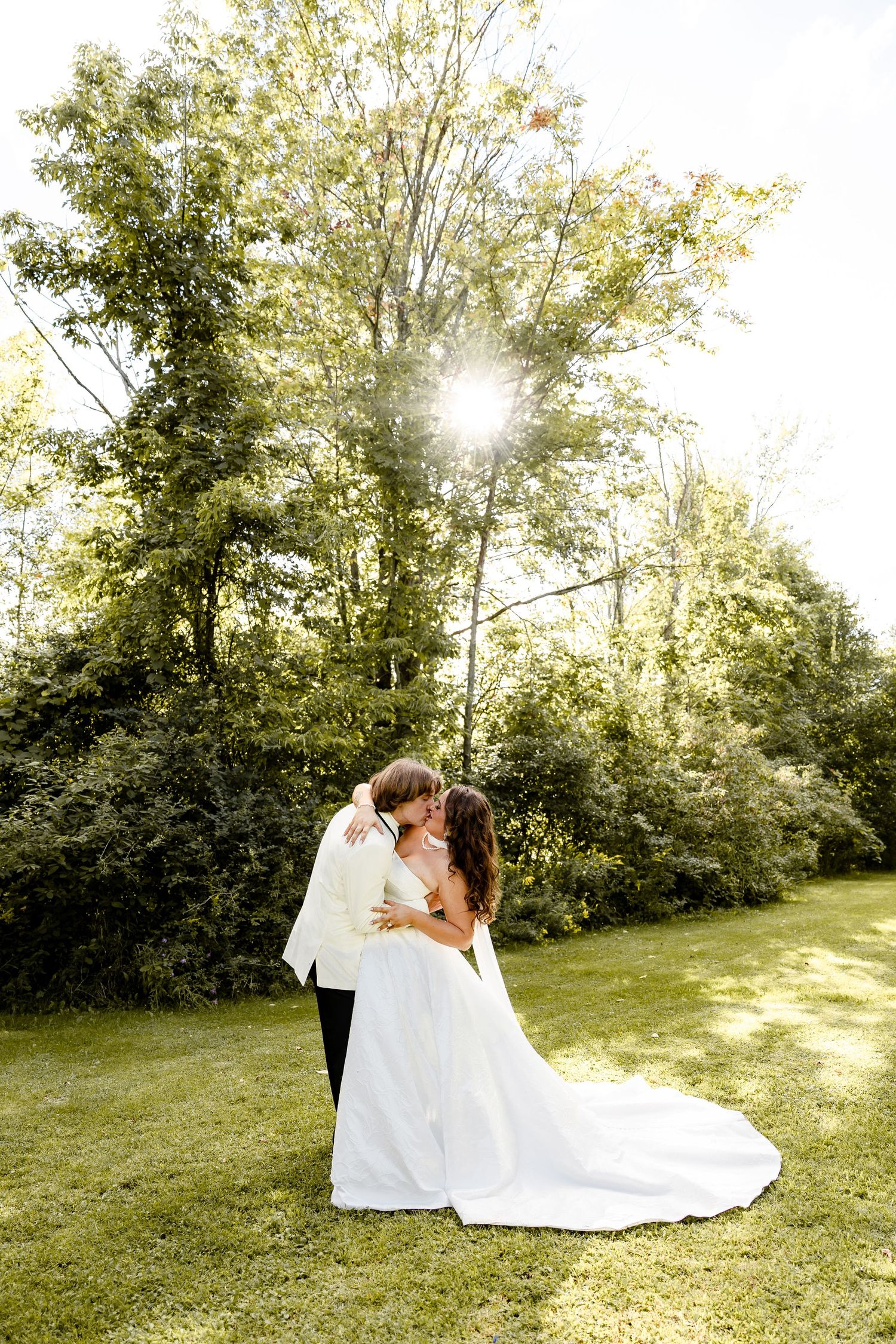 A bride and groom share a romantic kiss outdoors on green grass, surrounded by sunlit trees on their wedding day.