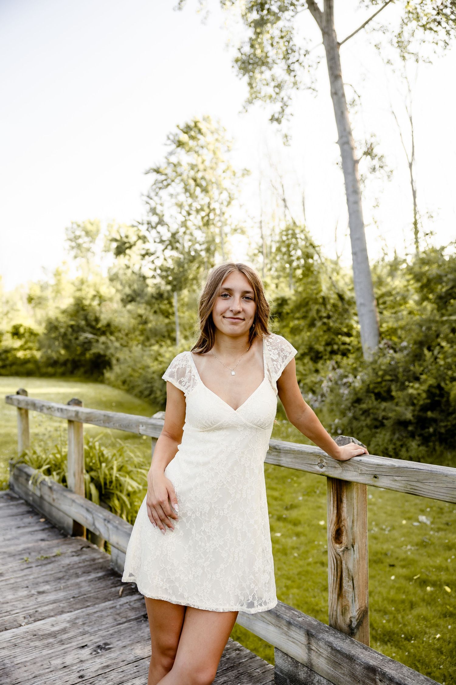 Young woman in white lace dress smiling on wooden bridge surrounded by lush green trees in golden sunlight.