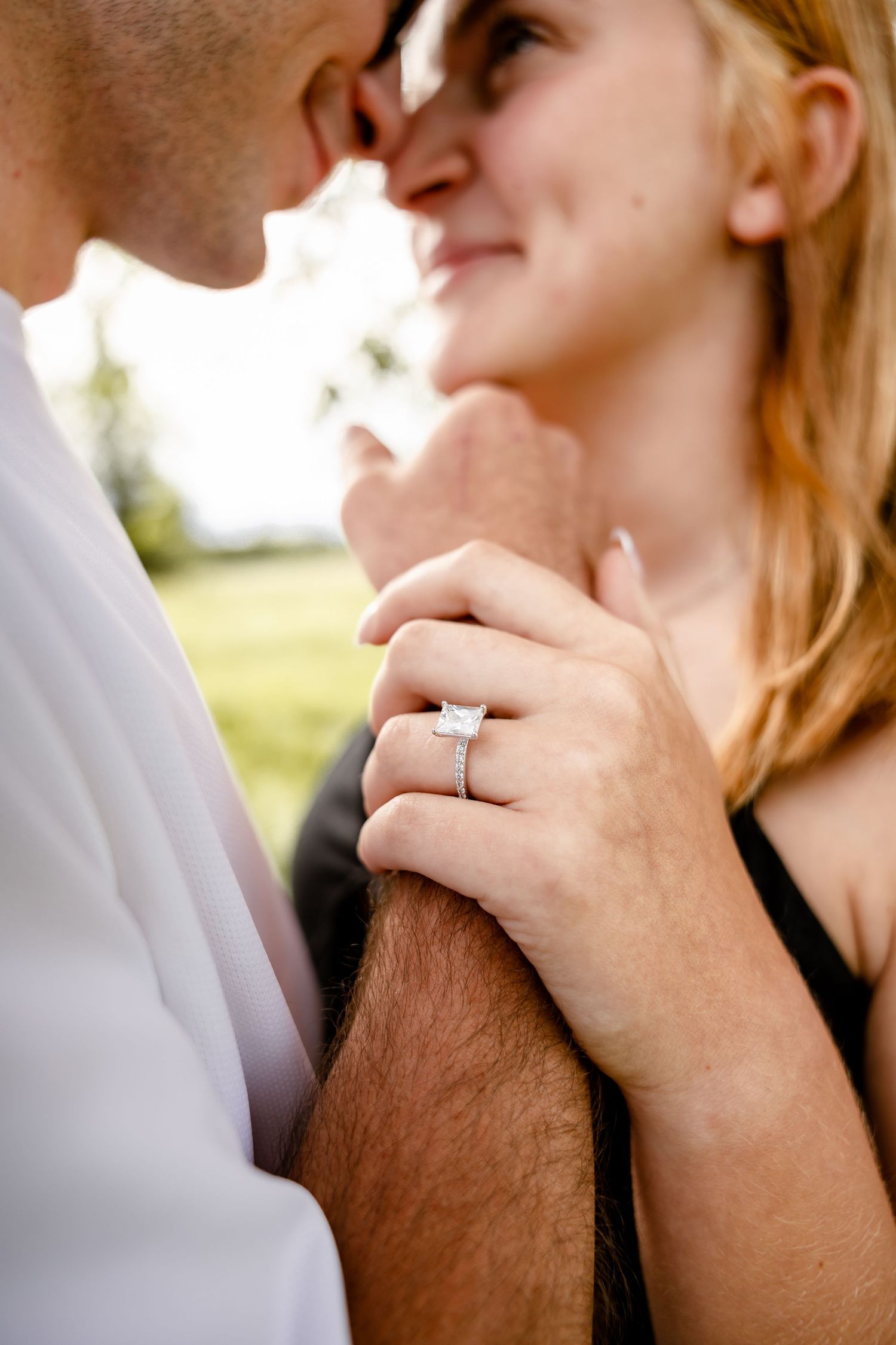 Couple nearly kissing outdoors, woman displaying diamond engagement ring on man's arm, green field background.
