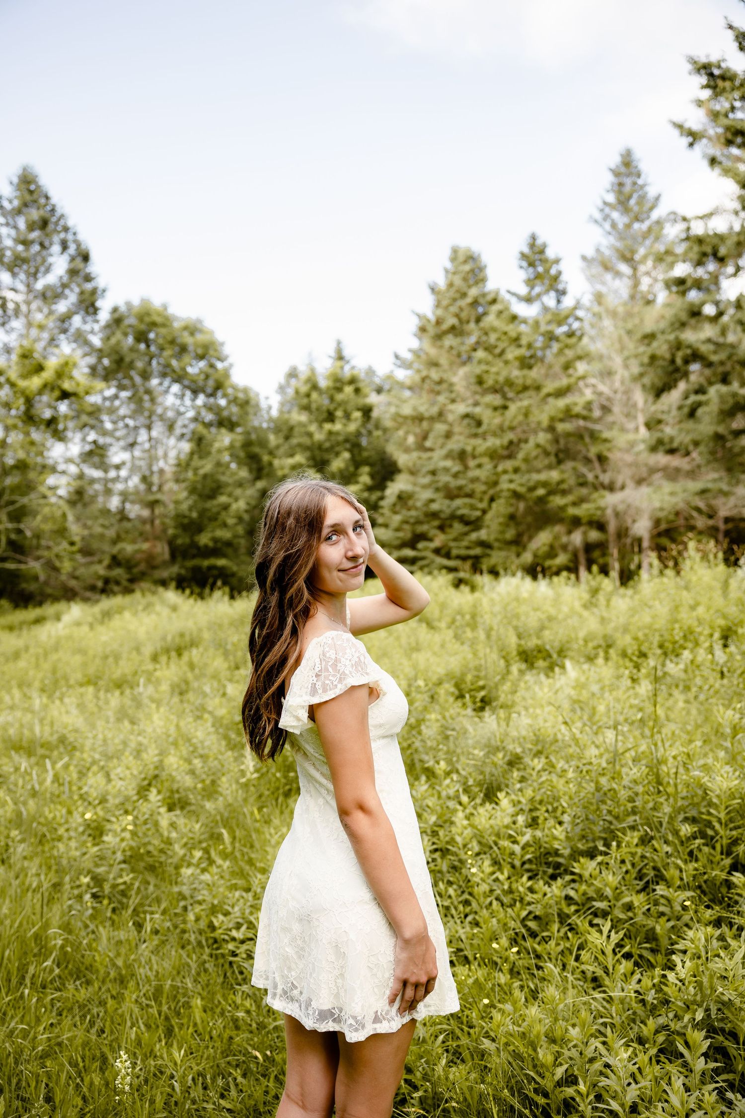 Young woman in white lace dress smiling in lush green meadow with pine trees in background.