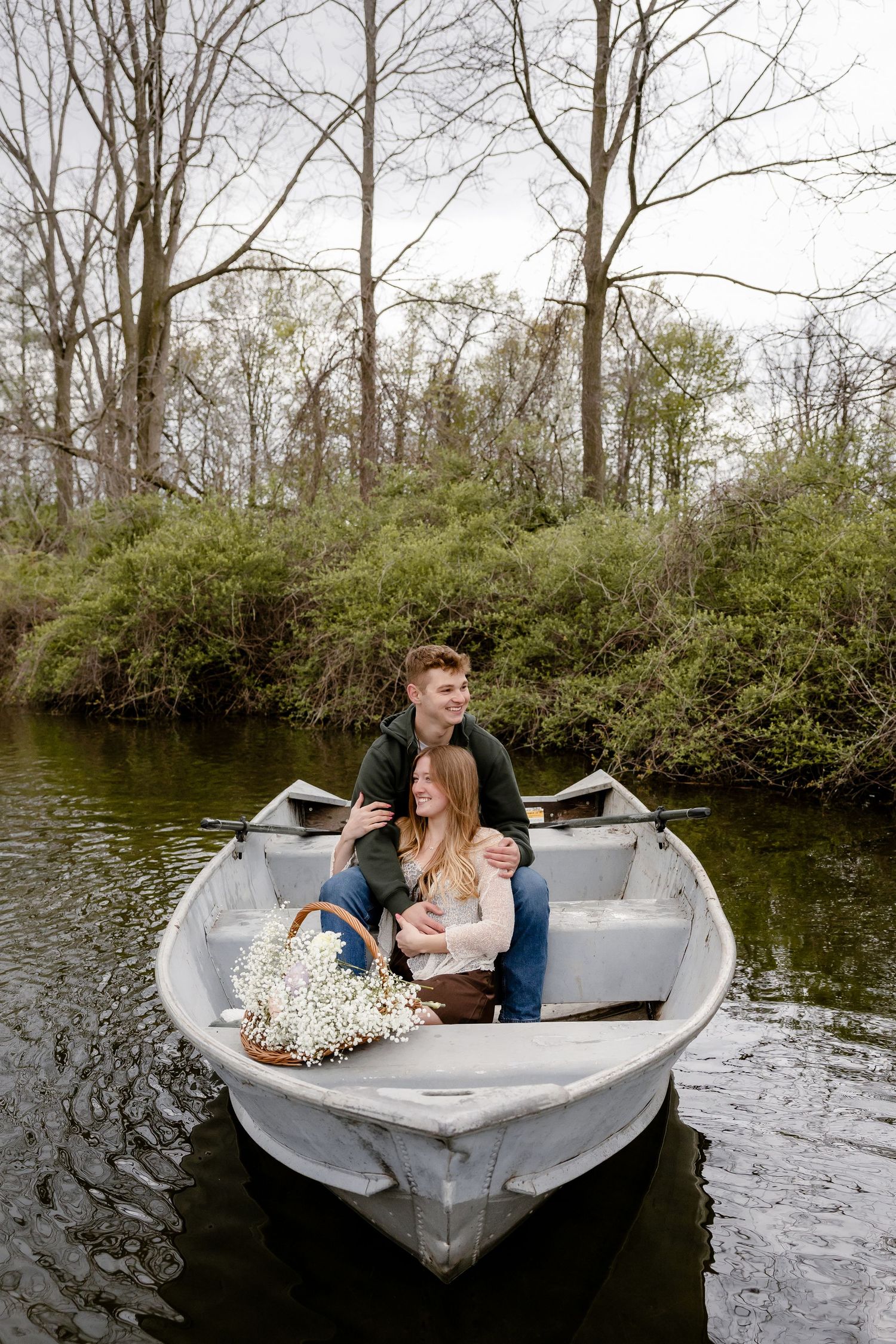 Romantic couple sitting together in a rowboat on a calm river surrounded by lush green trees, with a basket of white flowers.