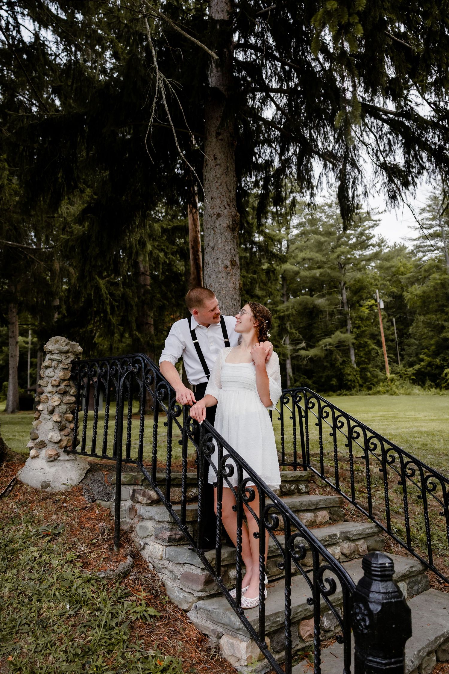 Bride and groom share a romantic moment on stone steps with black iron railings surrounded by lush green trees.