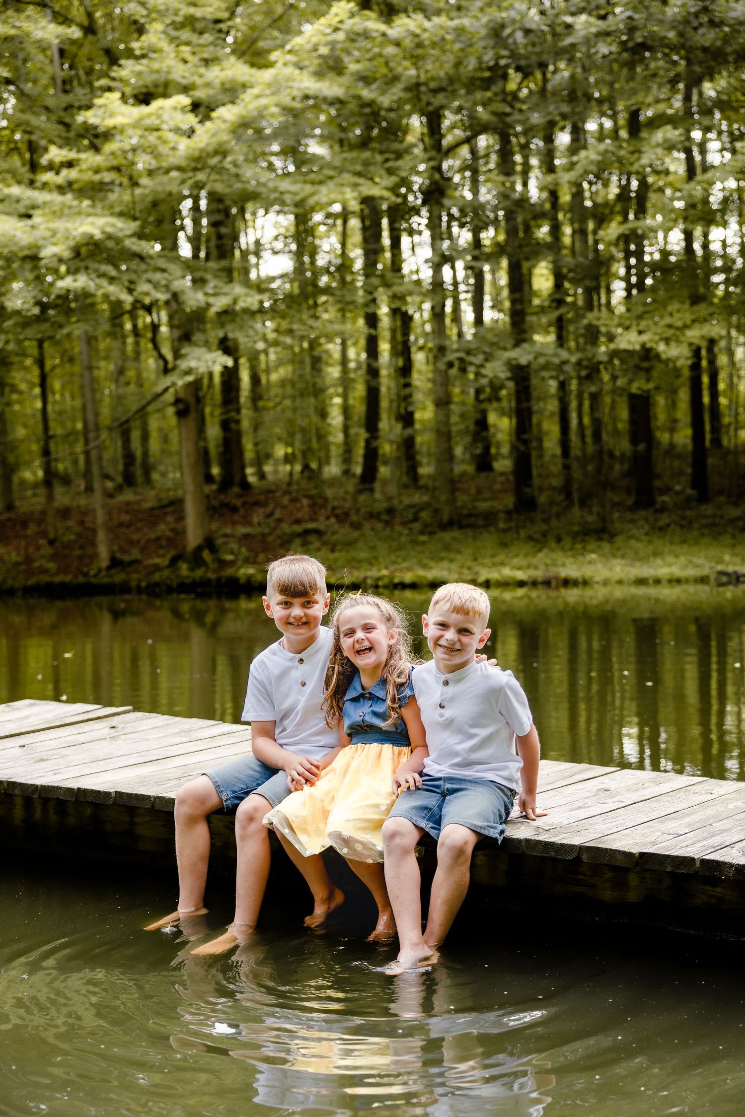 Three children sit barefoot on a wooden dock over a calm lake surrounded by lush green forest trees.