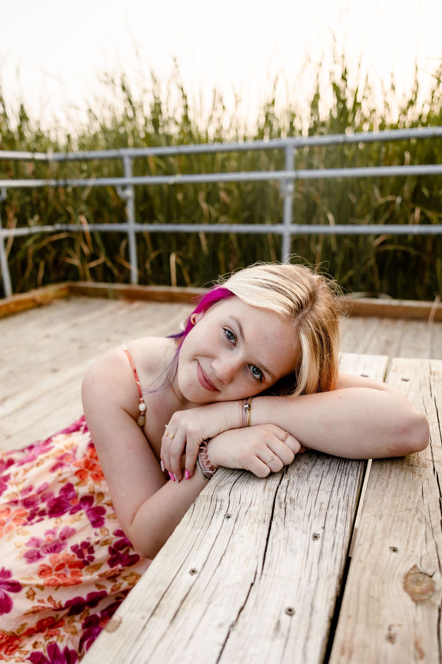 Young blonde girl in floral dress resting on wooden dock, smiling at camera with tall grass background.