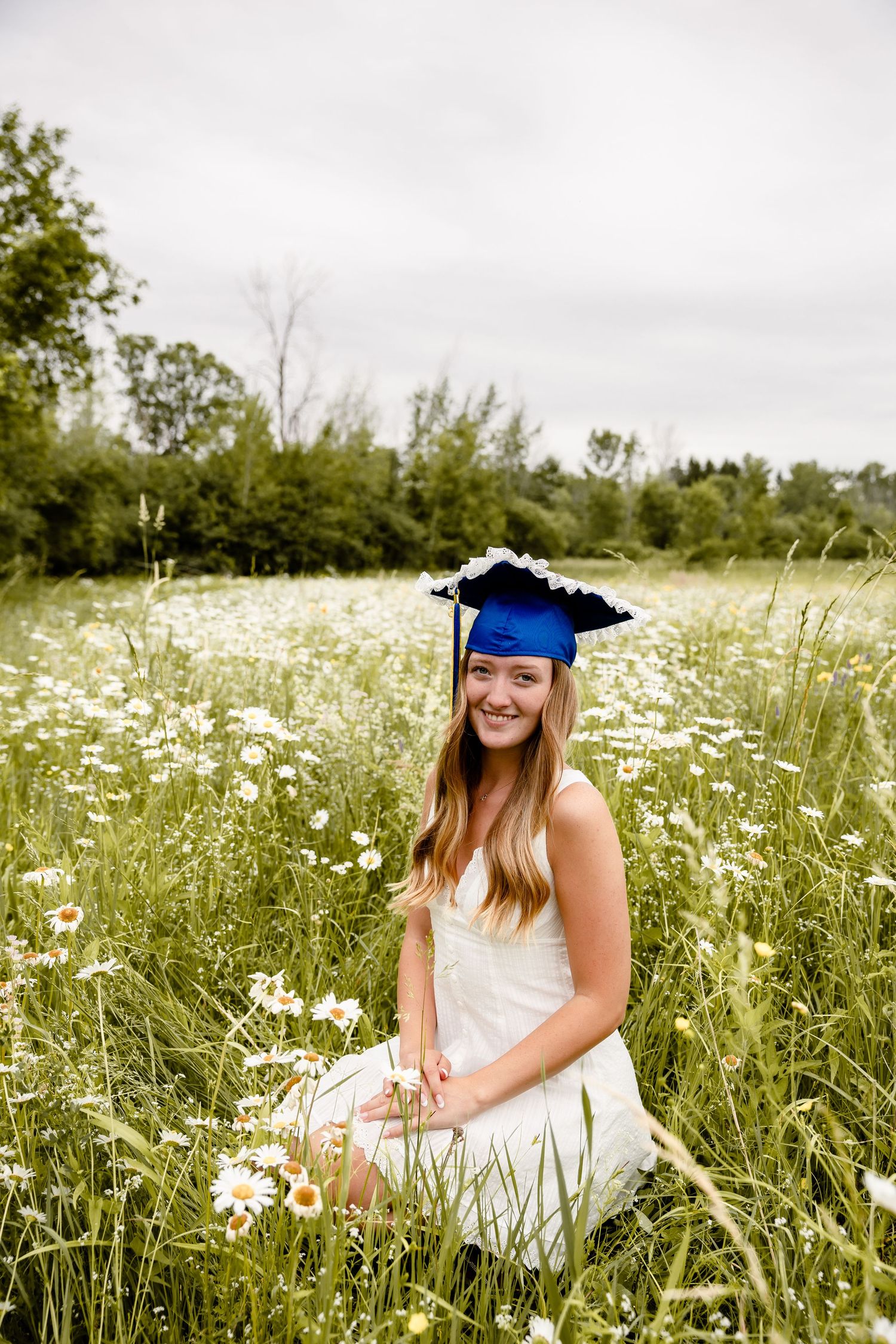 Smiling graduate in white dress and blue cap sitting in a field of white daisies on a cloudy day.
