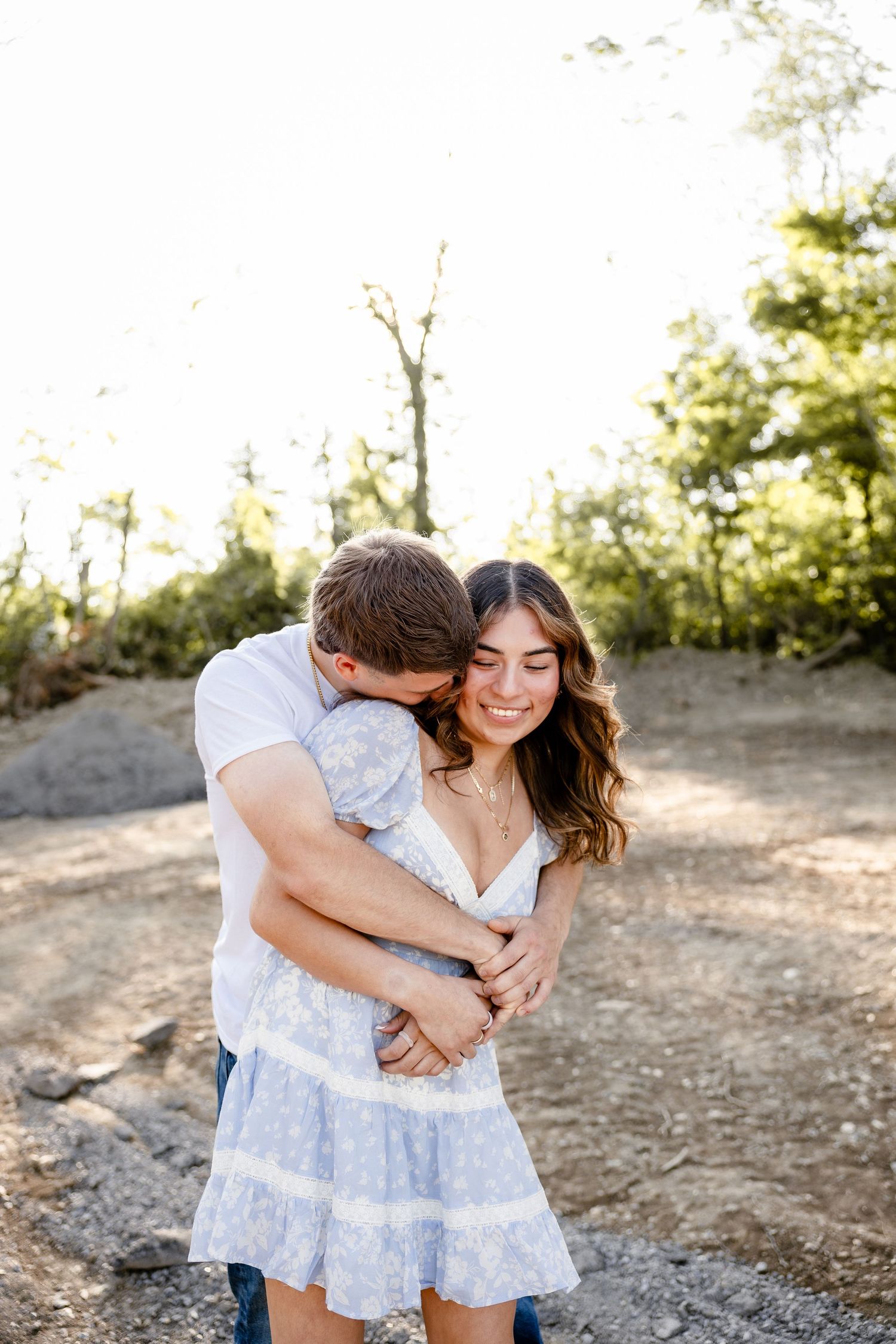 A couple embraces outdoors, man kissing woman's cheek as she smiles in a light blue dress with sunlit trees behind.