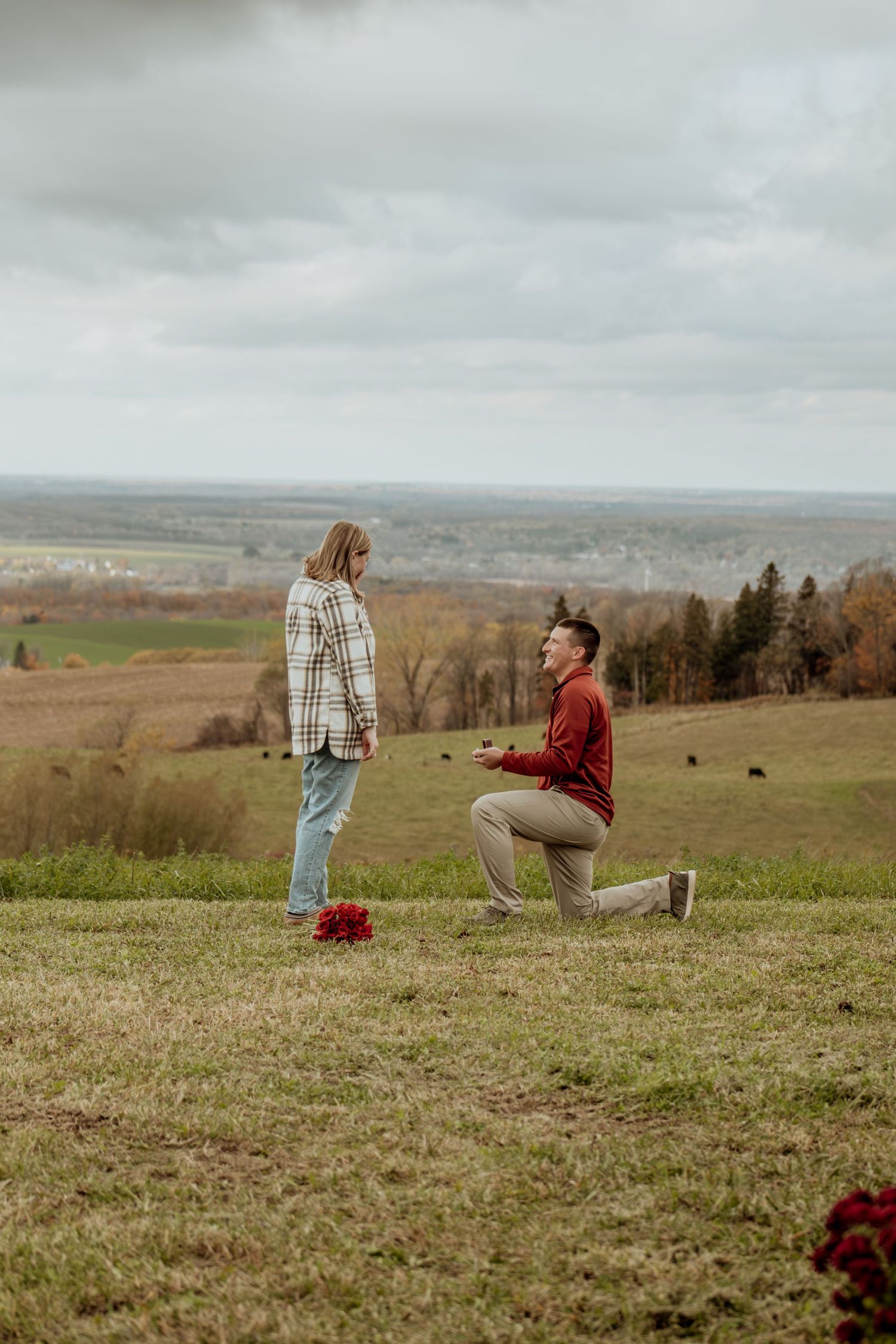 A couple stands in a scenic field overlooking rolling hills and farmland during a cloudy autumn day.