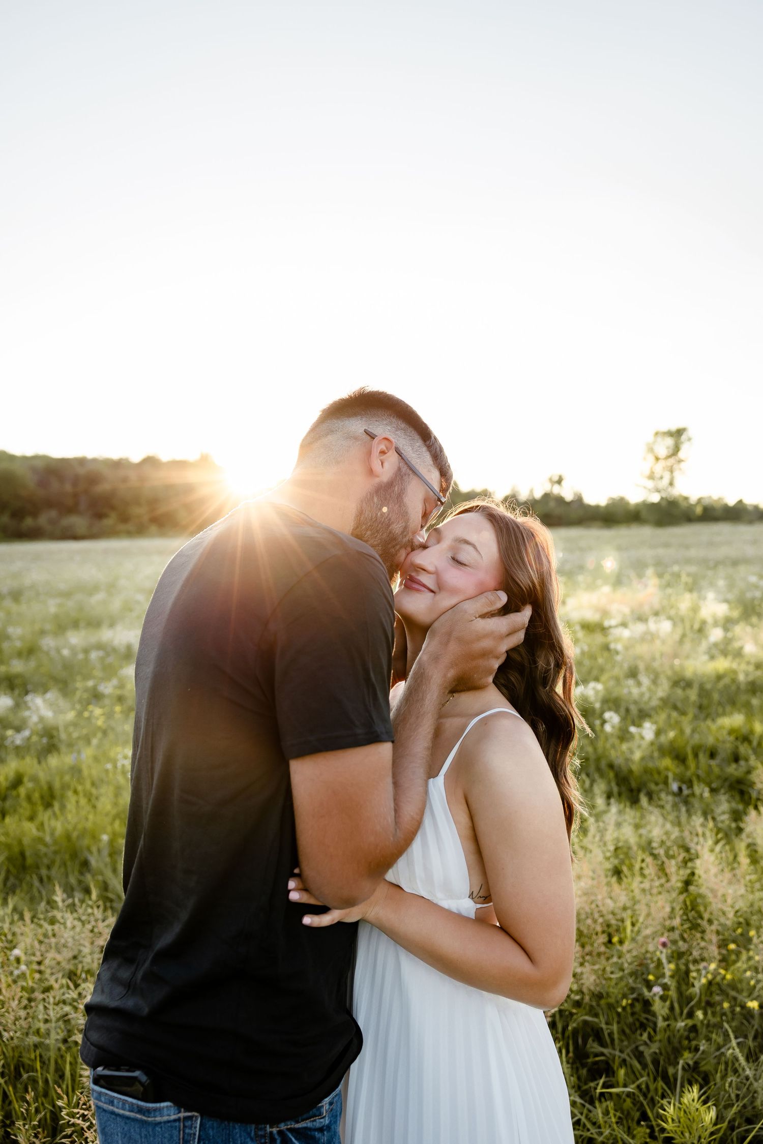 Couple sharing a romantic kiss in a sunlit field at golden hour, woman in white dress smiling joyfully.