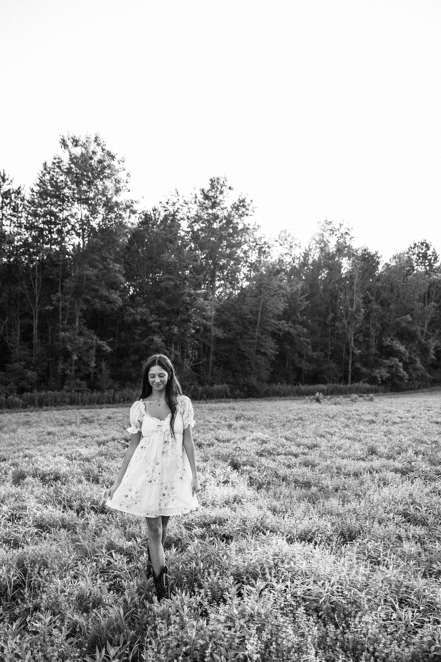 Black and white photo of a young woman in a floral dress walking through a field with trees in the background.