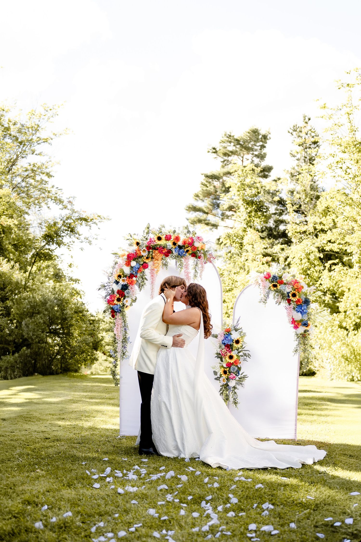 Bride and groom share first kiss under colorful floral arch on green lawn at outdoor wedding ceremony.