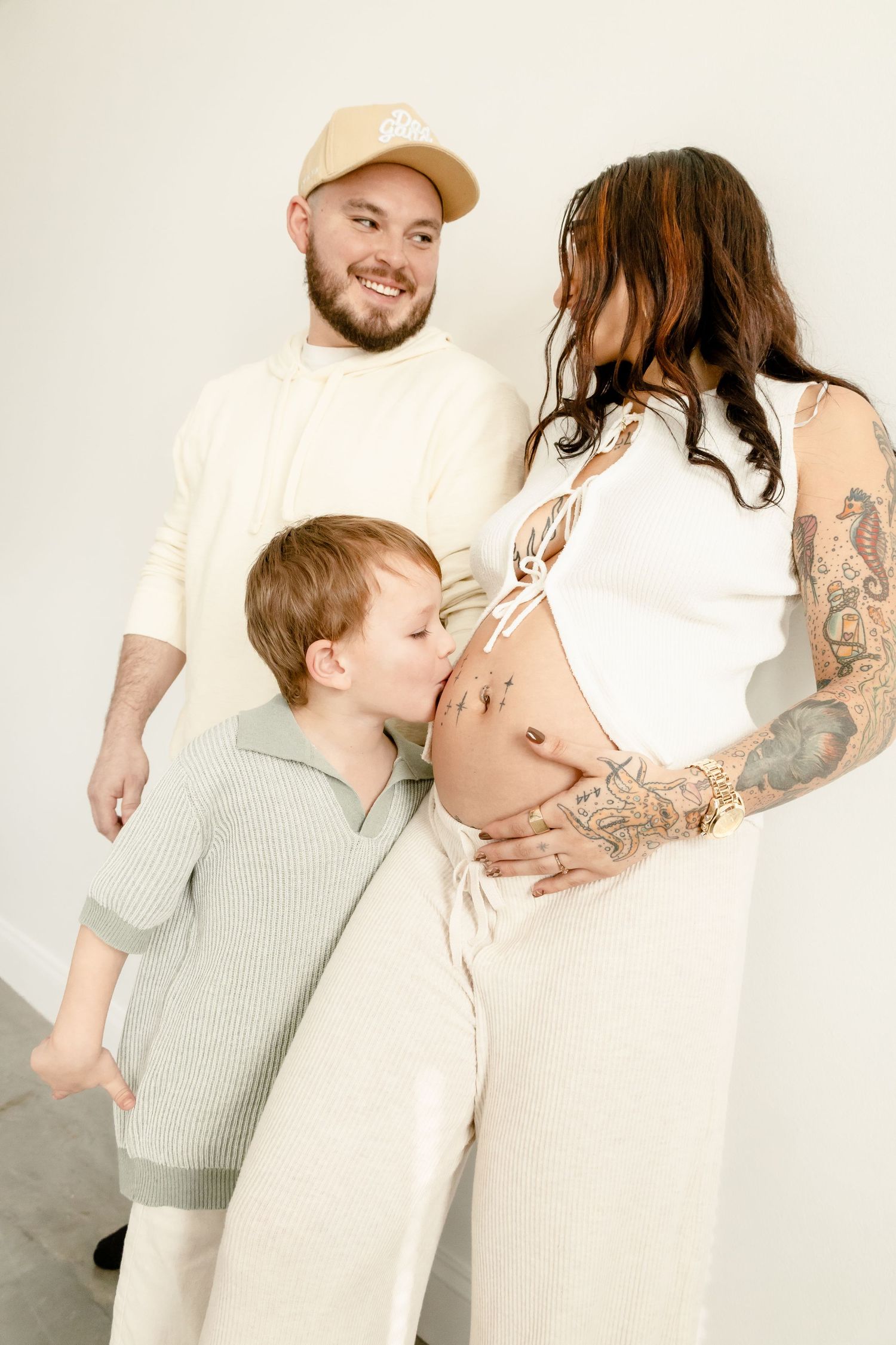 A young boy kisses his pregnant mother's belly while the father smiles warmly in a bright, neutral-toned family maternity photo.