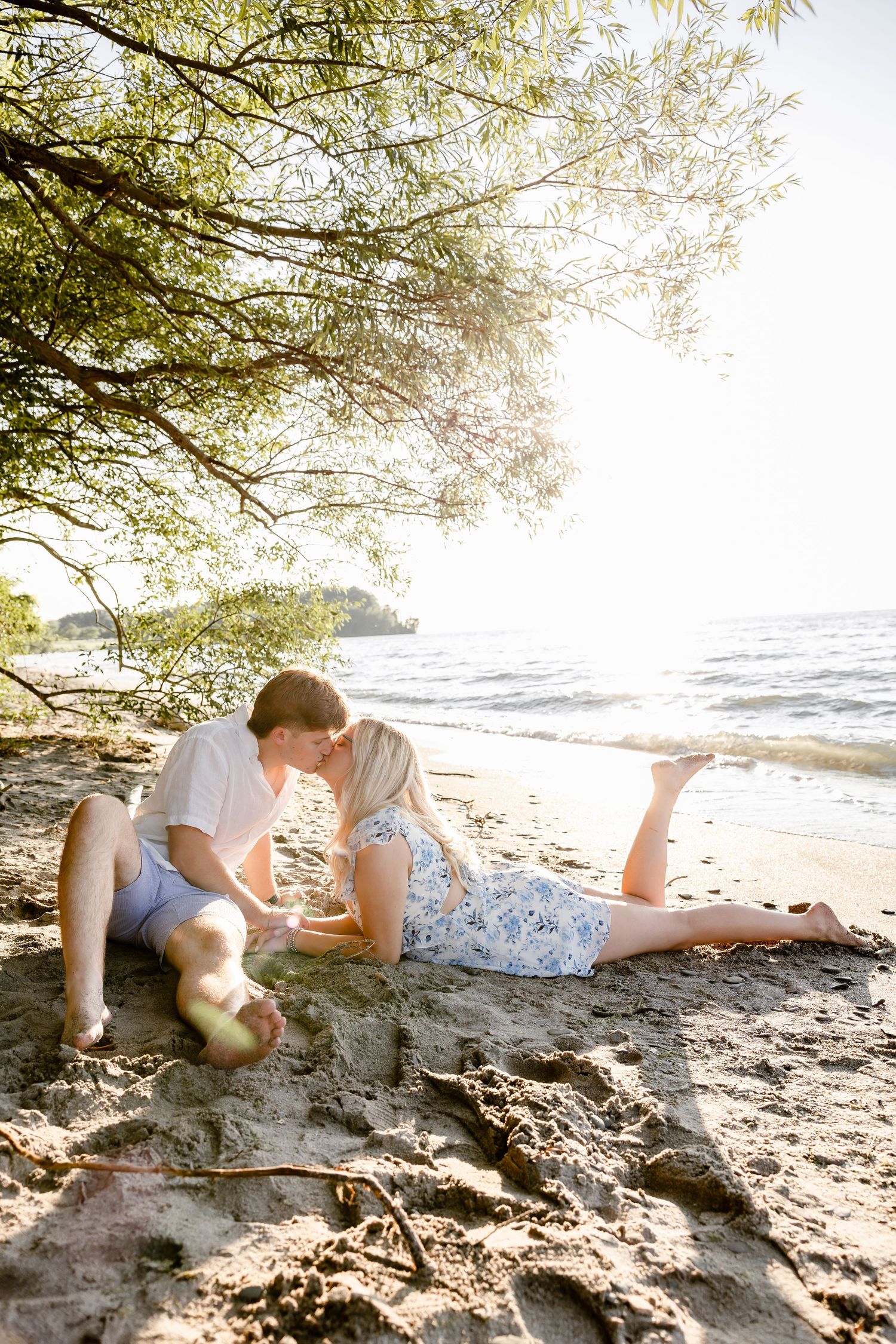 Romantic couple kissing on sandy beach shore under green tree with sunlit ocean backdrop.