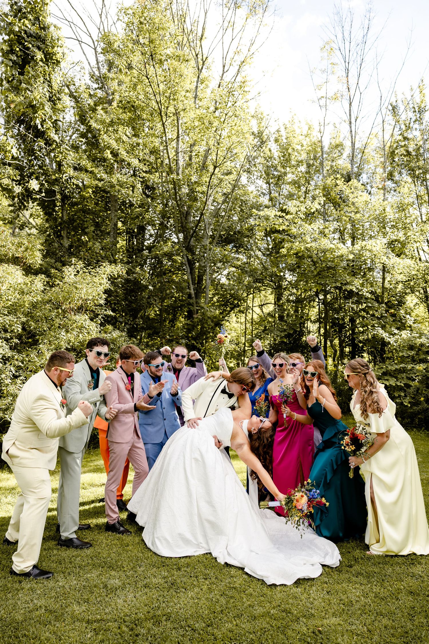 Wedding party in colorful attire celebrating outdoors, bride in white gown surrounded by guests on green lawn.