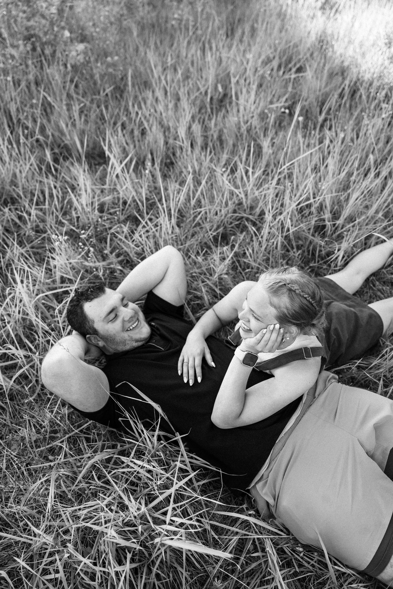 Black and white photo of a happy couple laughing together while lying in tall grass outdoors.