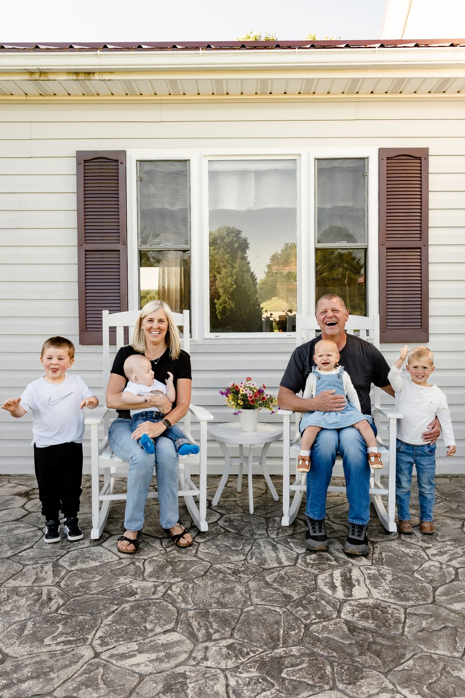 Family portrait on farmhouse porch with grandparents in rocking chairs holding babies and two young children standing beside them.