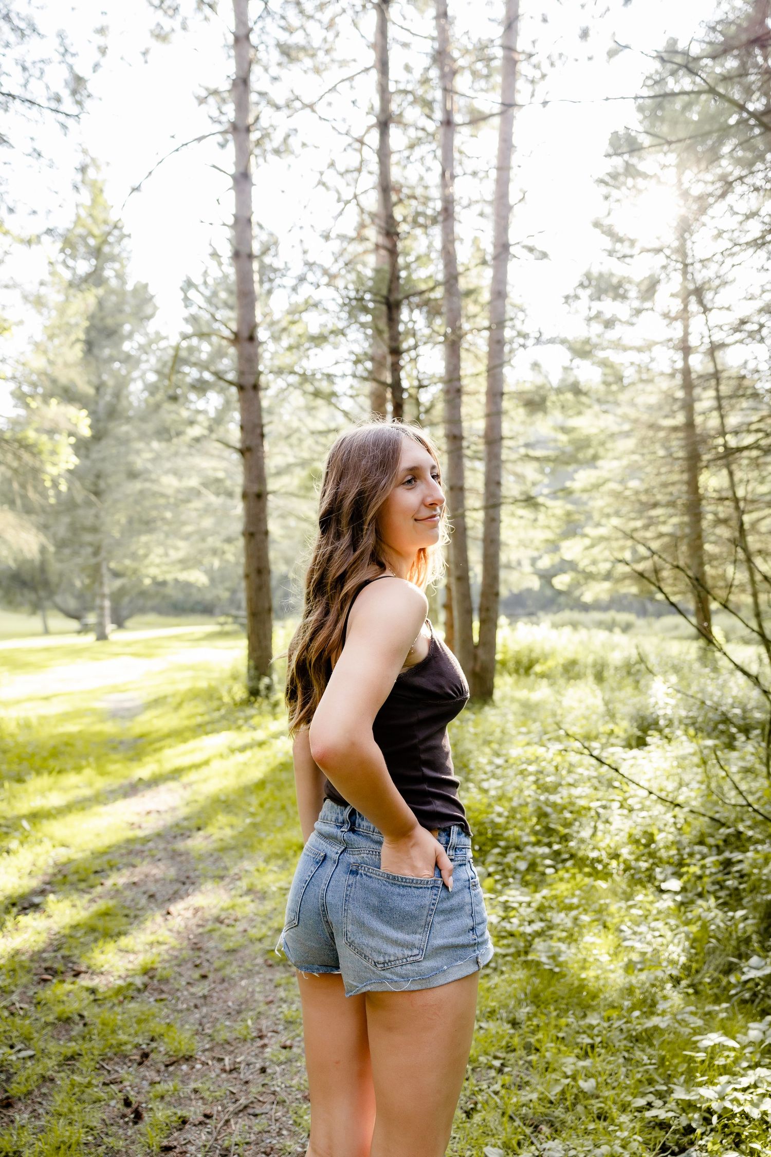 Young woman in black tank top and denim shorts smiling in a sunlit forest path surrounded by tall pine trees.