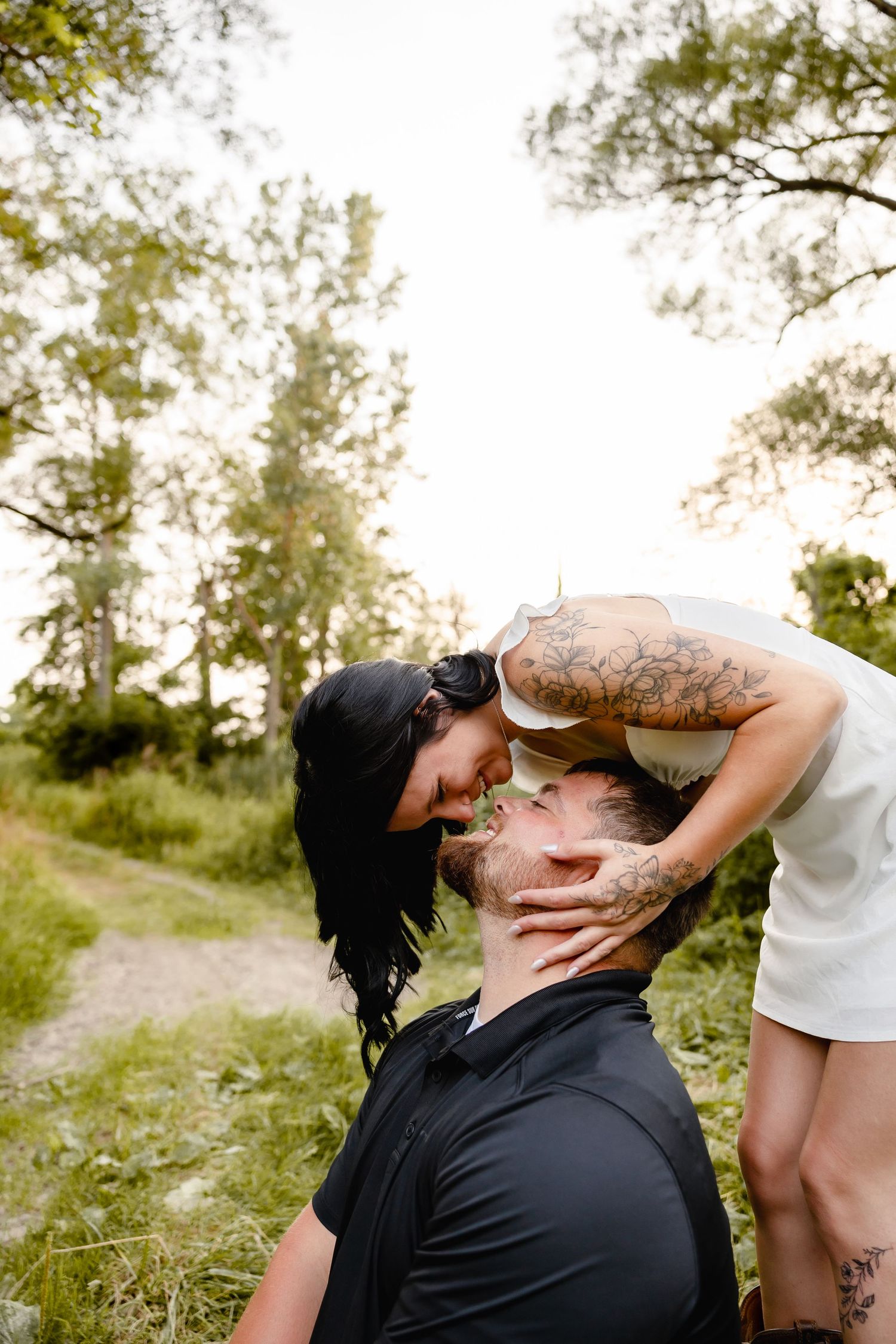 A tattooed couple shares a romantic moment outdoors, with the woman leaning down to kiss the mans forehead in a lush green setting.