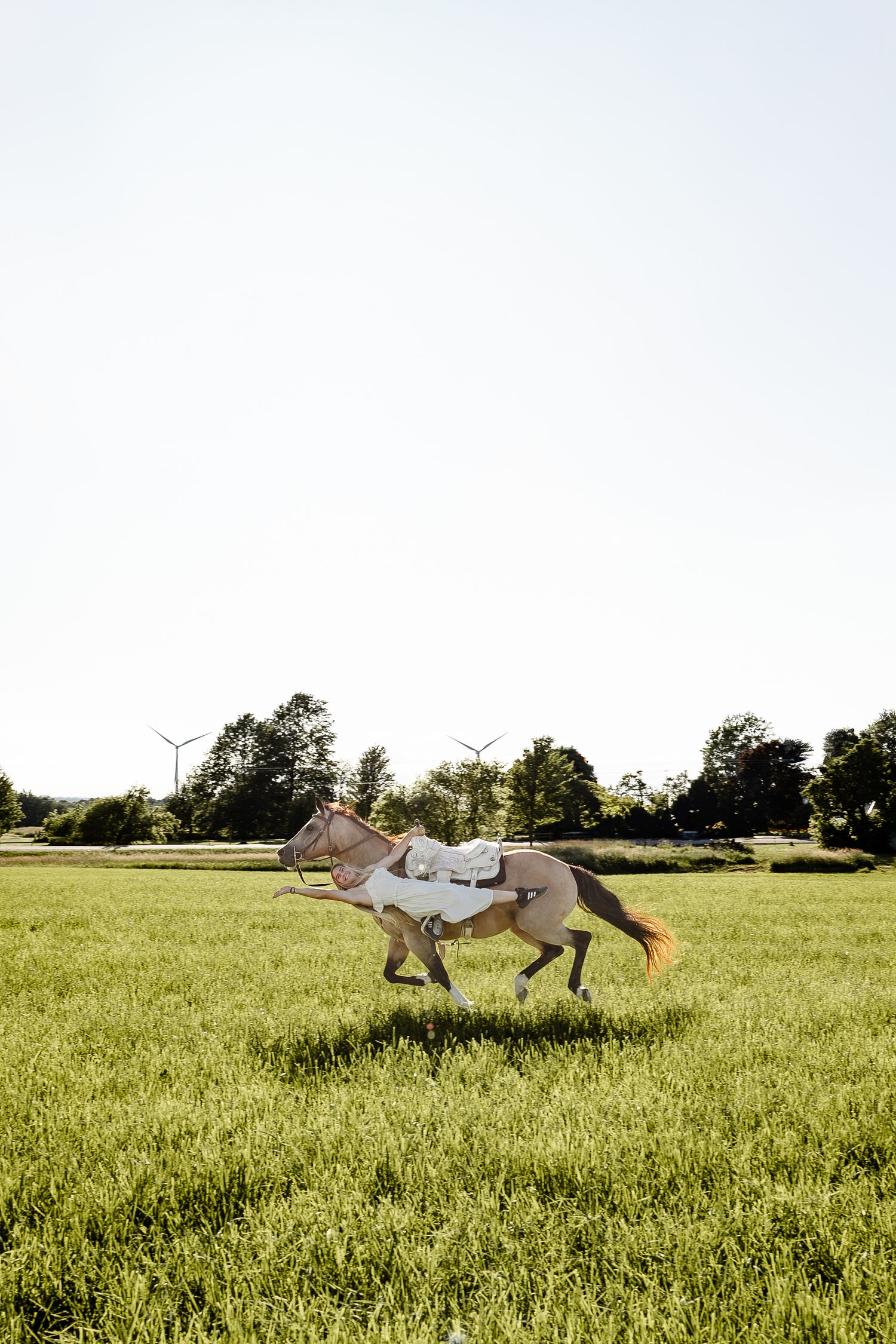 A horse galloping freely across a lush green field with wind turbines and trees in the background.