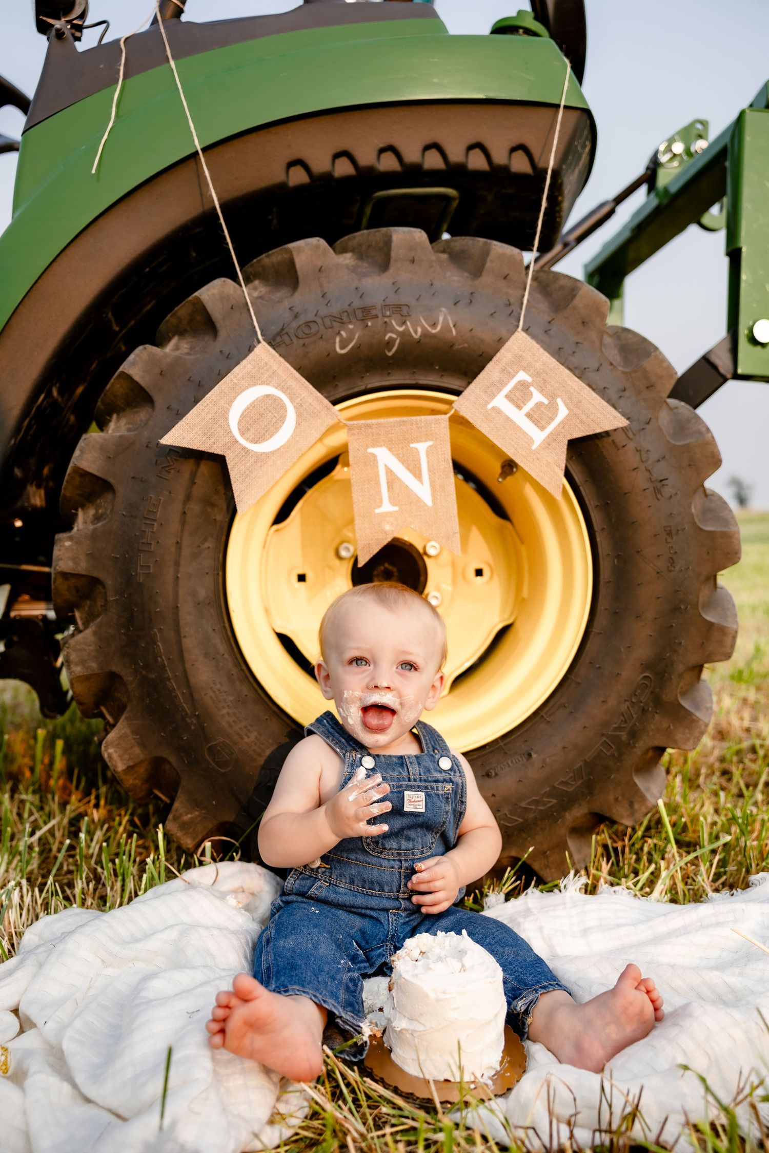 Baby in overalls sits smiling in front of John Deere tractor tire with ONE birthday banner for first birthday cake smash.