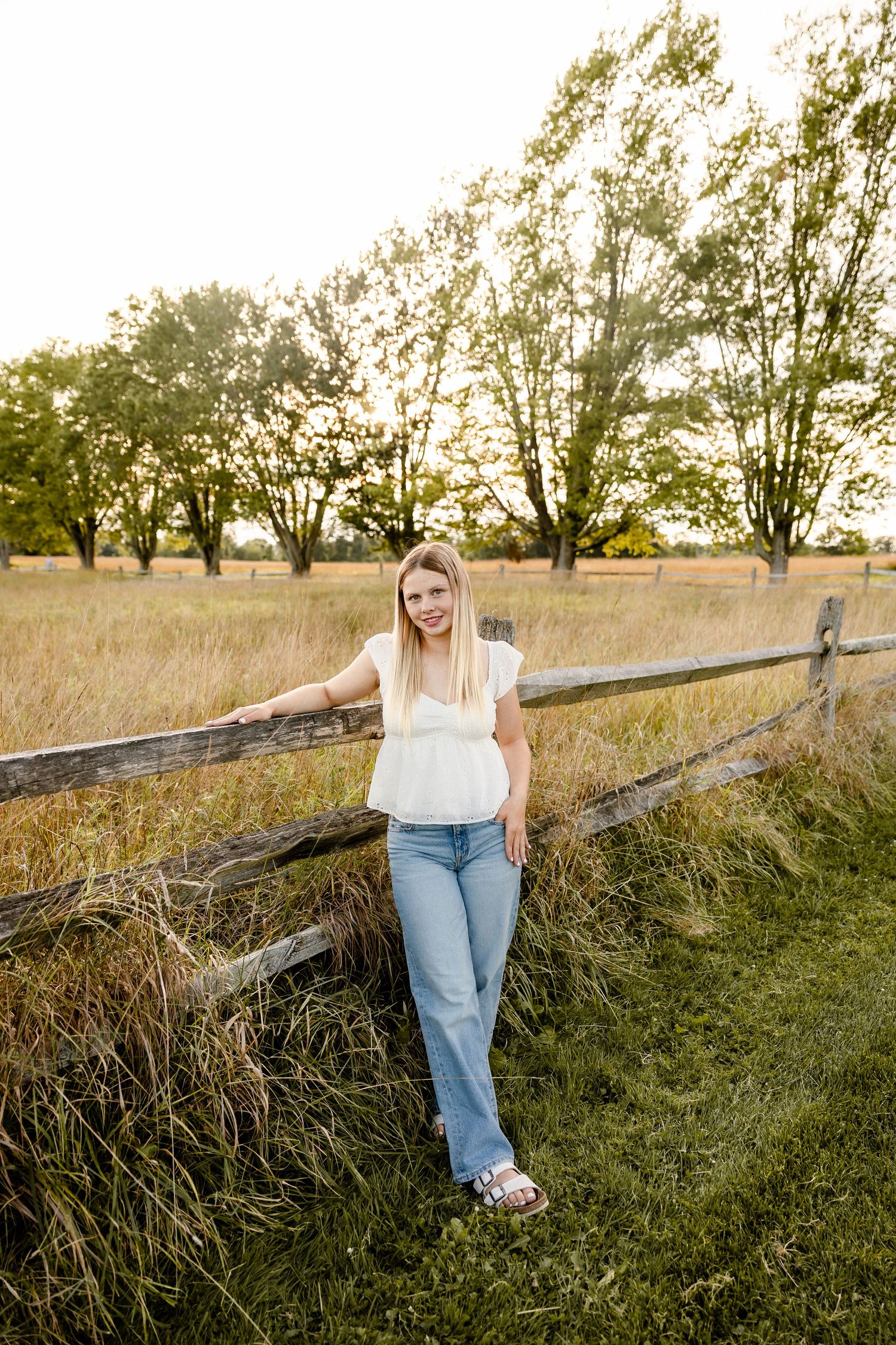 Smiling blonde woman in white top and jeans leaning against rustic wooden fence in golden field.