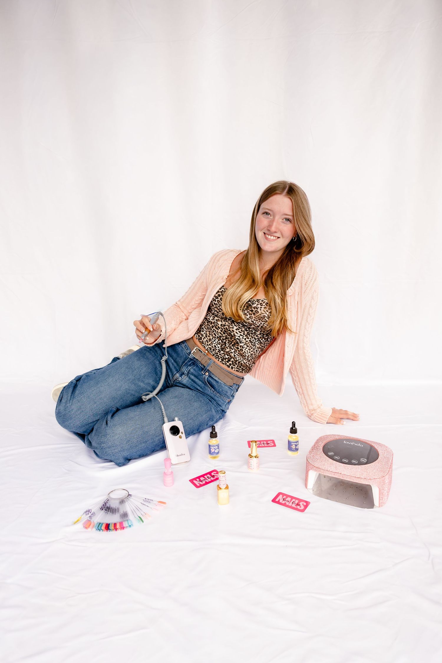 Young woman sitting on white backdrop with gel nail kit, UV lamp, nail polish, and manicure tools spread around her.
