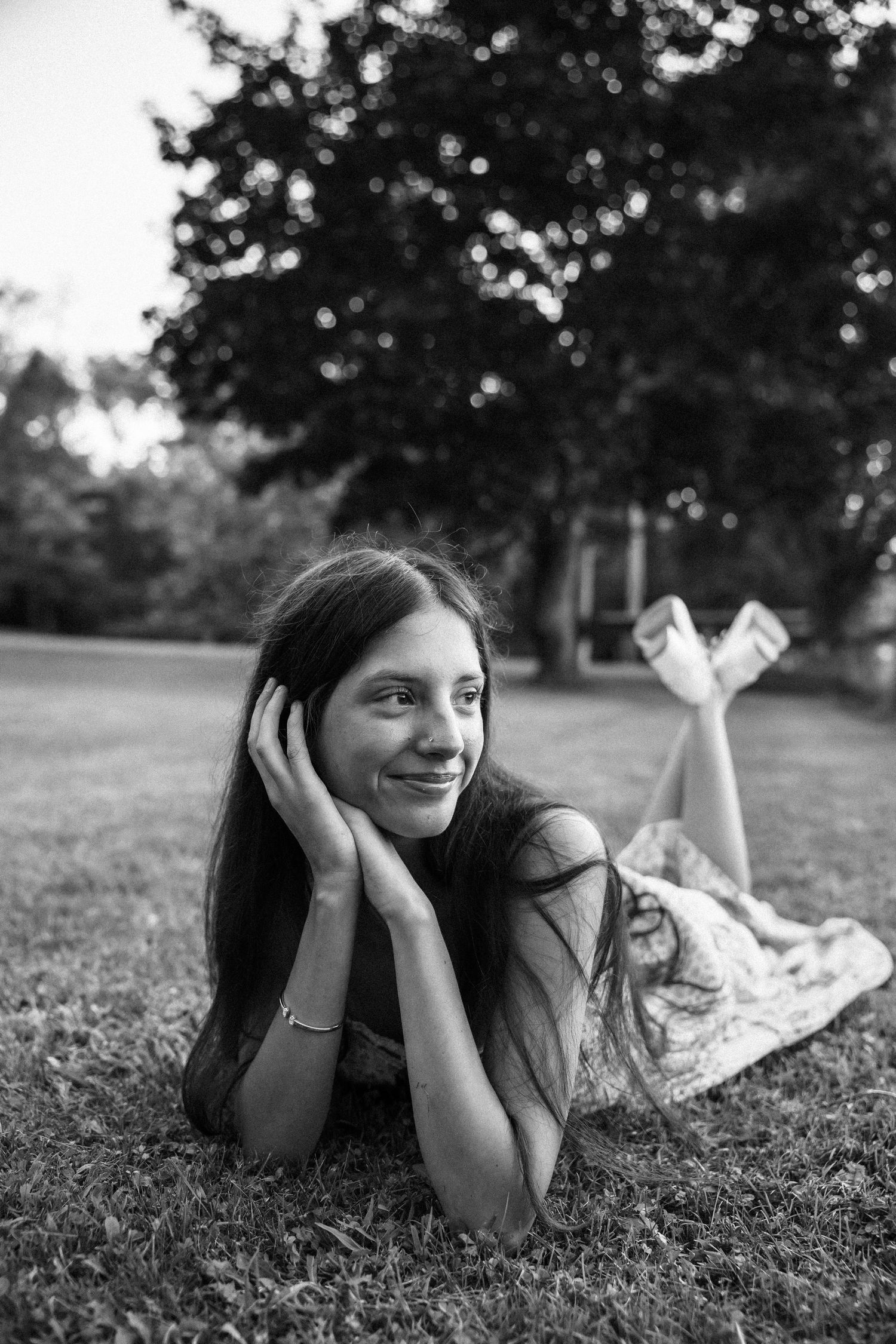 Black and white photo of a smiling young woman lying on grass in a park, resting her chin on her hands.